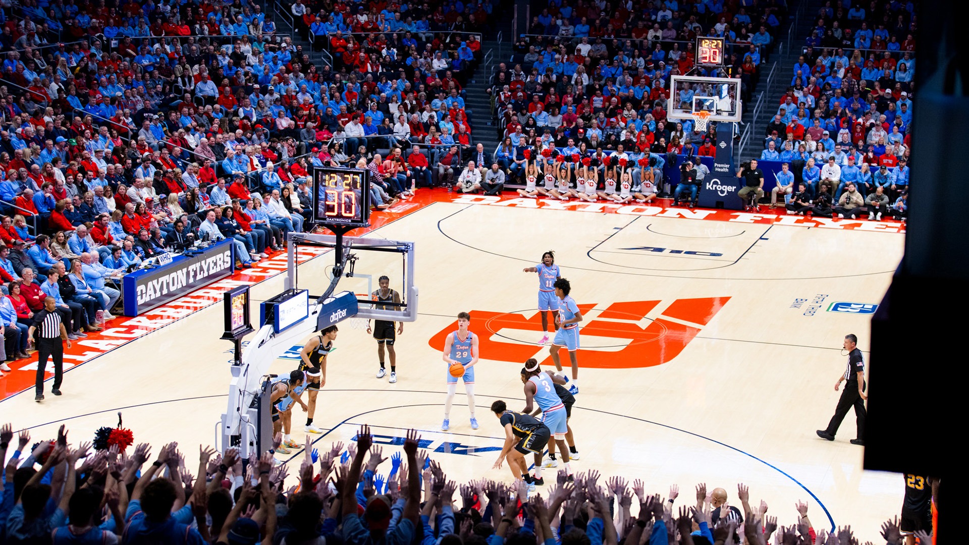 A Dayton Flyers basketball player prepares to shoot a free throw during a home game at UD Arena. The crowd, dressed mostly in red and blue, raises their hands behind the basket to distract the opposing team. Other players line the key, and the Dayton bench is visible in the background along with the court's large red 