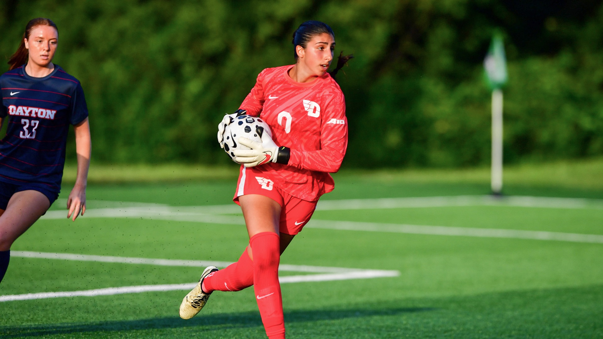 Dayton goalkeeper Batoul Reda, wearing a red uniform and gloves, holds the soccer ball securely while preparing to distribute it upfield during a match. A teammate in a navy Dayton jersey runs nearby on the turf field.