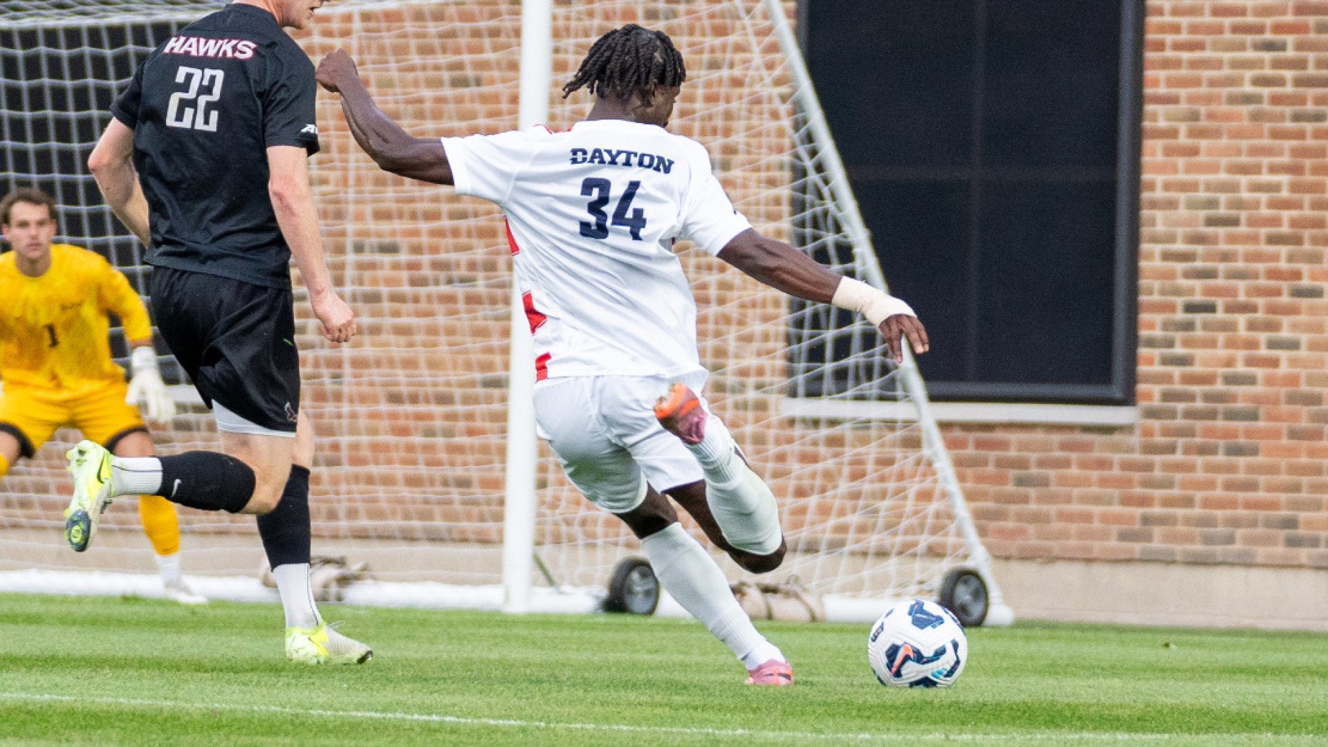 Men's soccer player Felix Buabeng, wearing a white jersey with the word DAYTON and the number 34, about to take a shot on goal with a defender on his left during a home game at Baujan Field