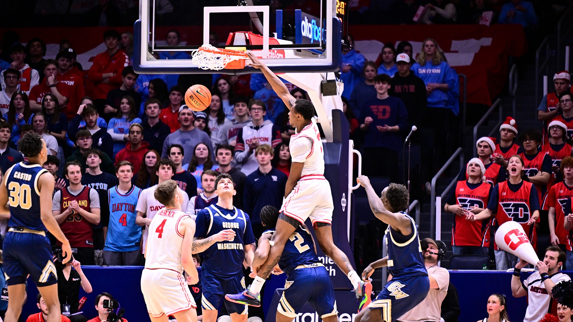 Dayton men's basketball player Keonte Jones, wearing a white uniform with the number seven, dunks a basketball over a George Washington defender.