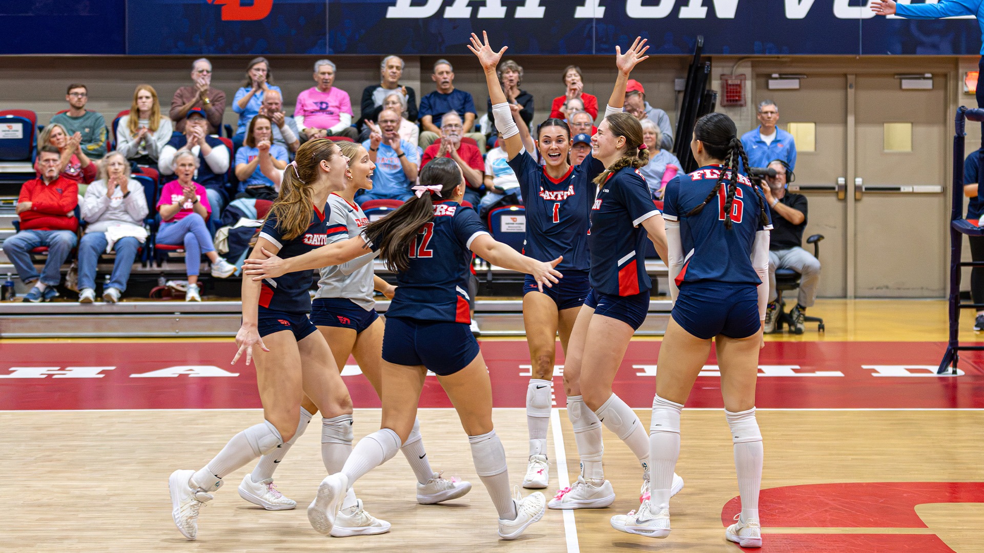 Volleyball players celebrate on the court after a point. They are wearing navy uniforms.