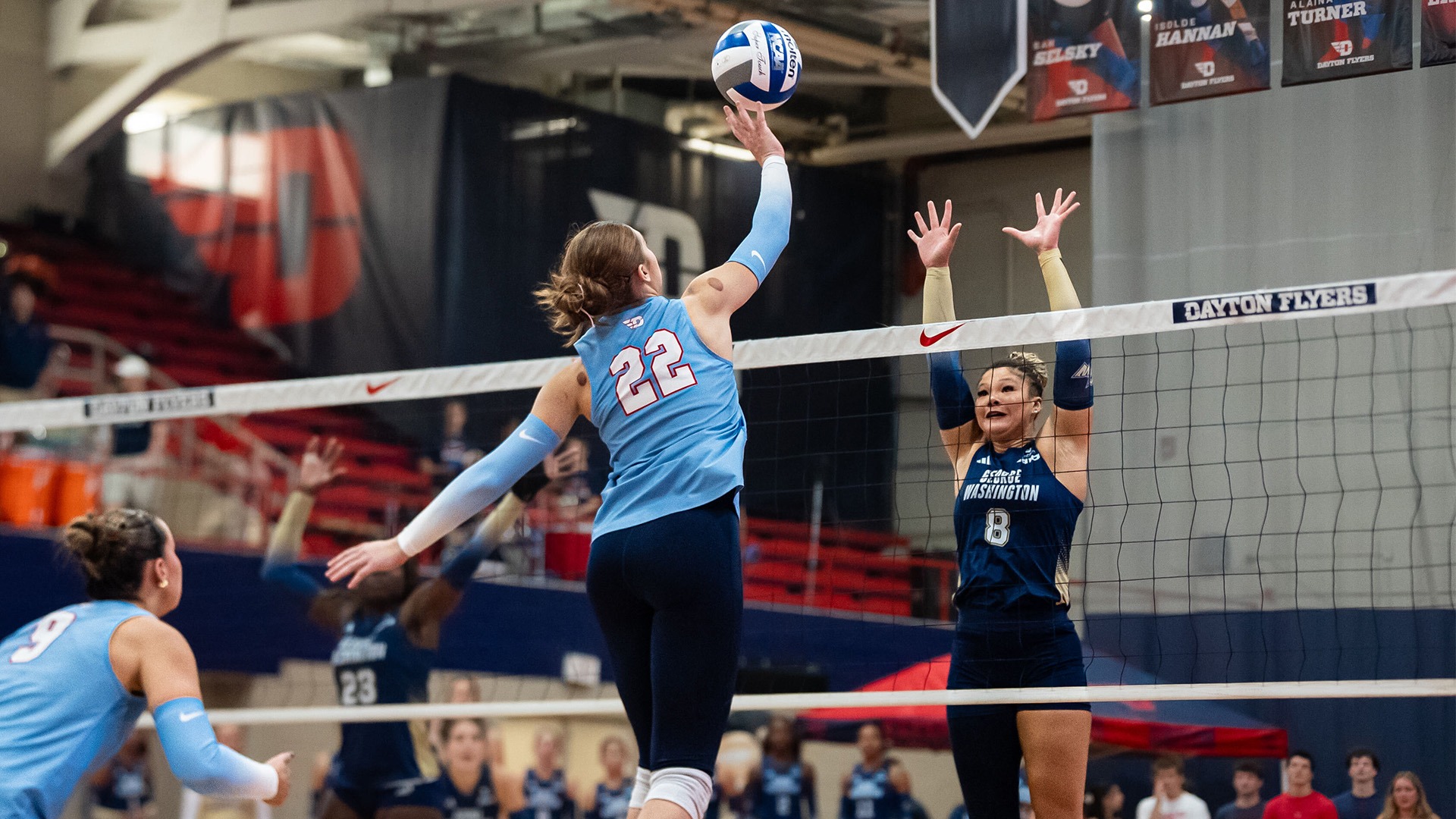 Sidney Sprada is in the air, attacking the ball at the net, while a George Washington player attempts to block the ball. She is wearing a chapel blue jersey and navy leggings with her back to the camera.