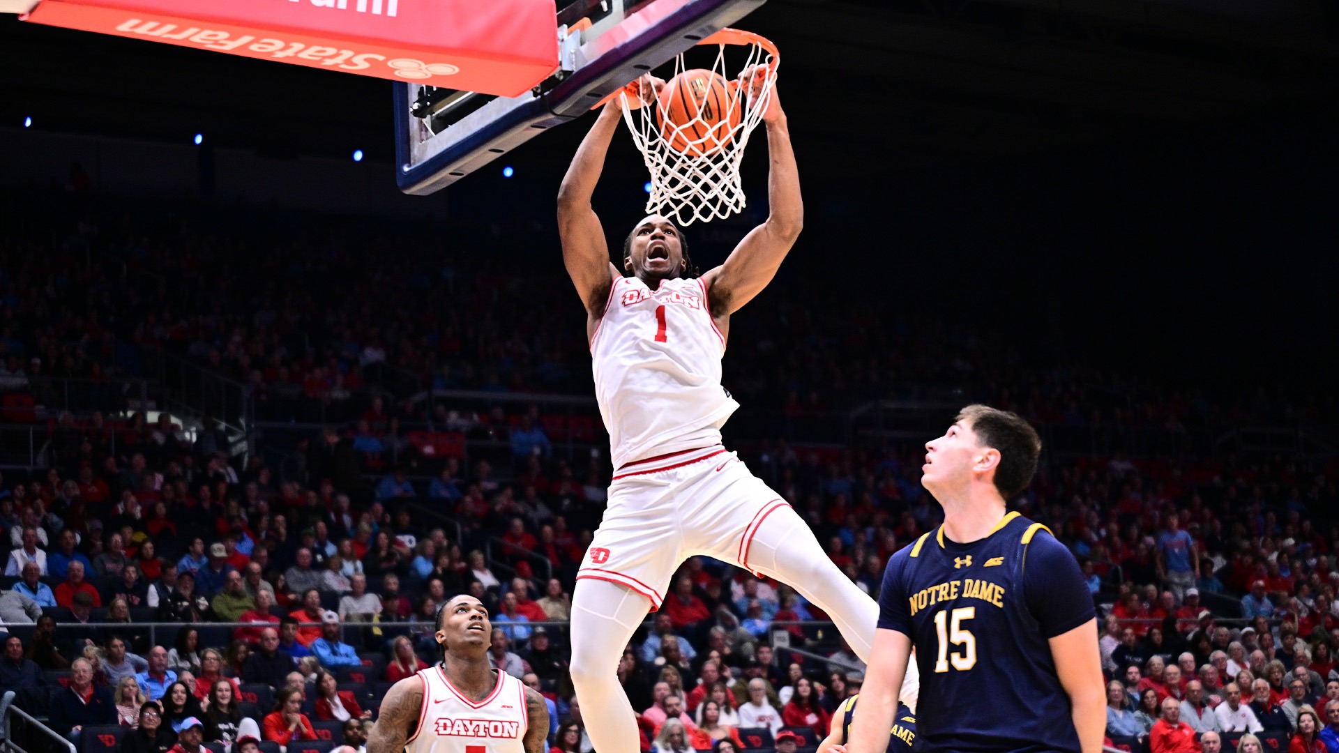 Malcolm Thomas holds onto the rim as the ball goes through the hoop. He is wearing a white uniform.