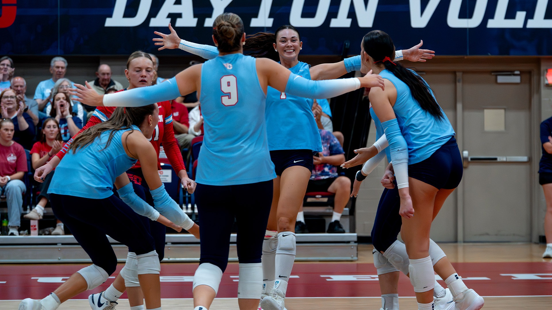 Volleyball players huddle in celebration after a point. They are wearing chapel blue uniforms and navy shorts.