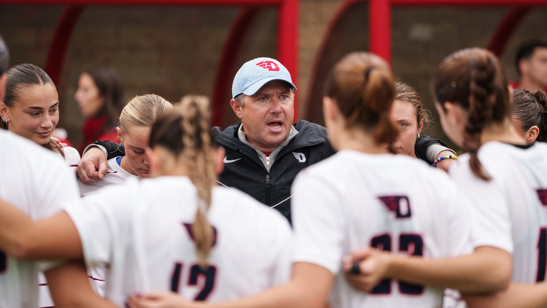 Dayton women's soccer coach Eric Golz motivates his team in the pre-game huddle while wearing a black jacket and chapel blue Dayton hat.