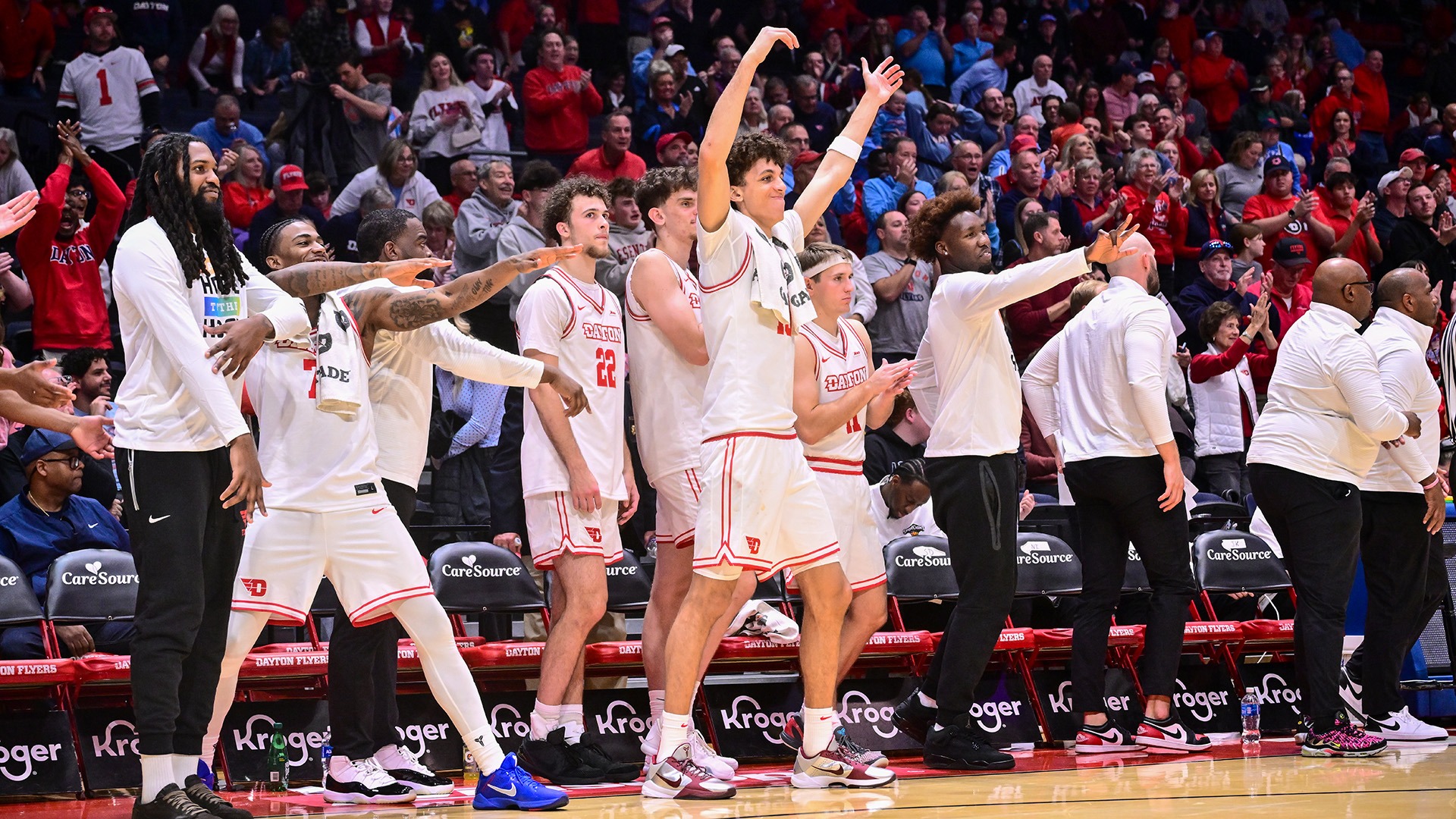Dayton men’s basketball players and coaches celebrate on the sideline with arms raised and extended as fans cheer and applaud in the stands at UD Arena.