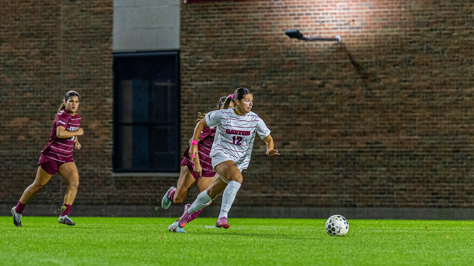 Dayton women's soccer player Lauryn Contini sprints toward the ball while pursued by two Fordham defenders during a nighttime match.