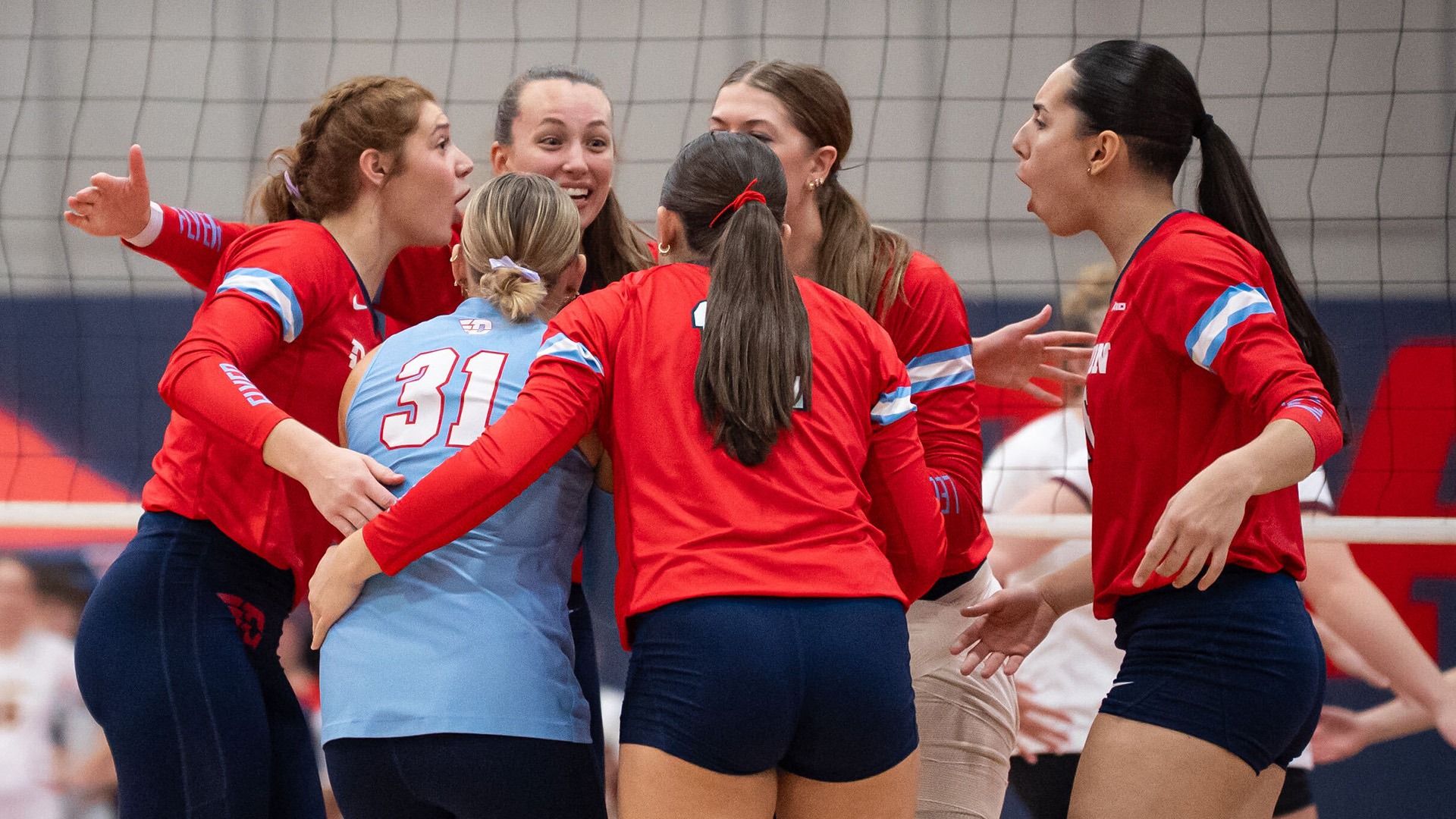 Volleyball players huddle in front of the net after a point. They are wearing red uniforms, while the libero is in a chapel blue jersey. All are in navy pants and shorts.