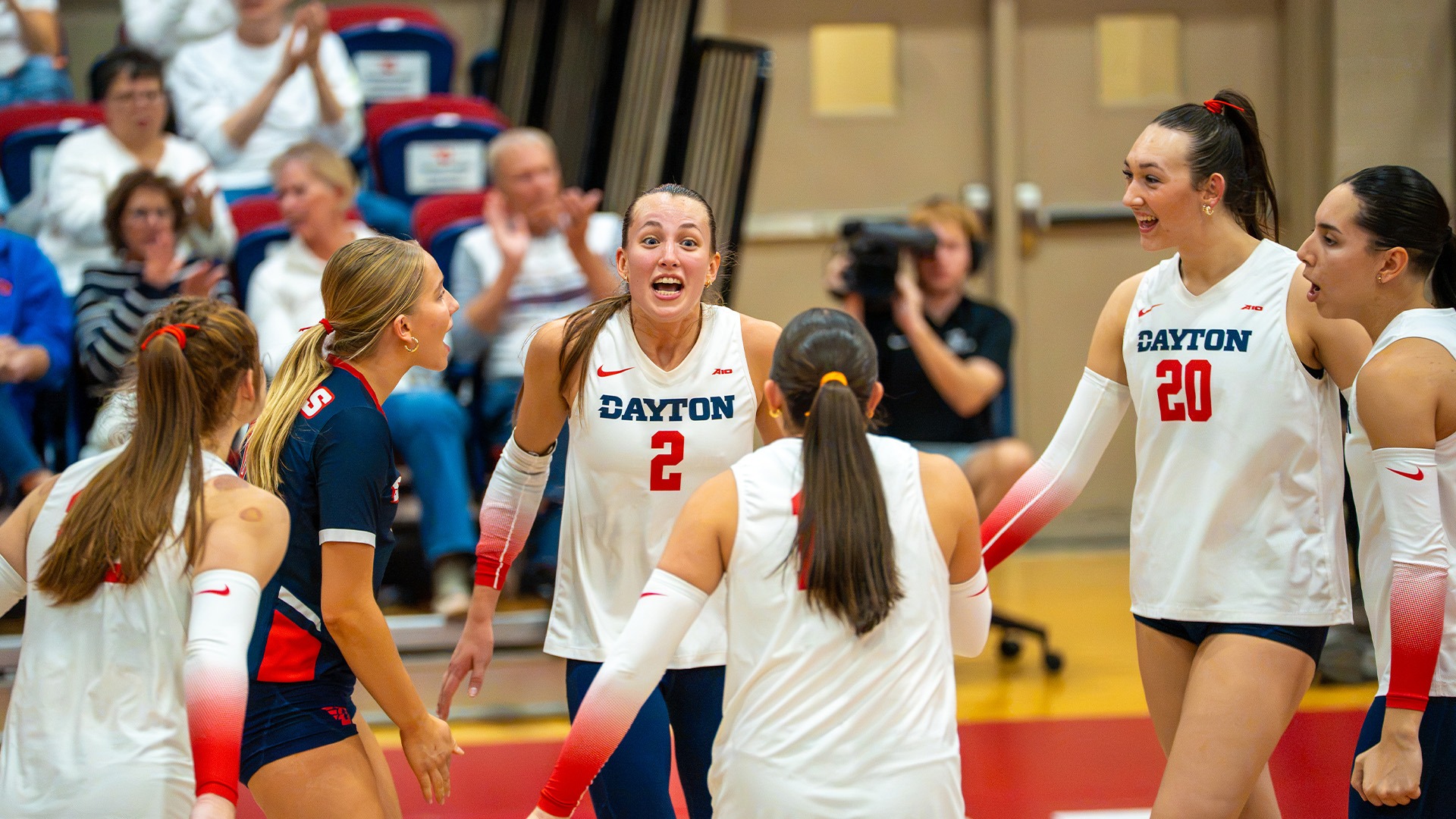 Volleyball players celebrate on the court after a point. Players are wearing white jerseys with navy bottoms, while the libero is in all navy.