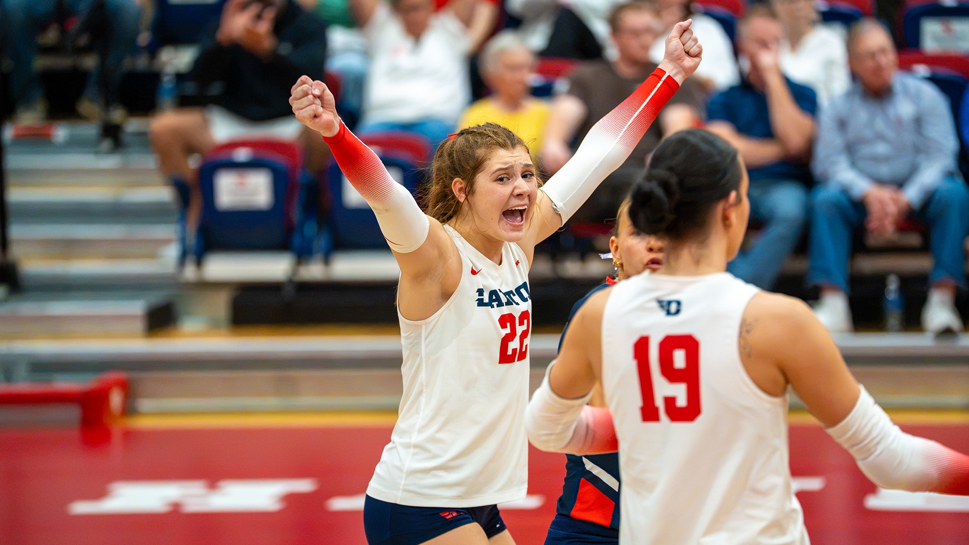 Sidney Sprada looks toward the bench with her arms up, yelling in celebration after a point. She is wearing a white jersey.