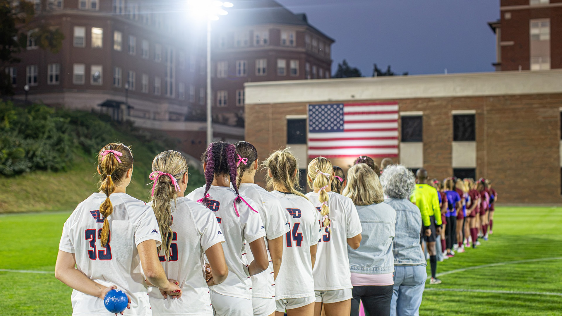 Dayton women’s soccer players and staff stand along the sideline facing an American flag during a pregame ceremony at a lighted soccer field.