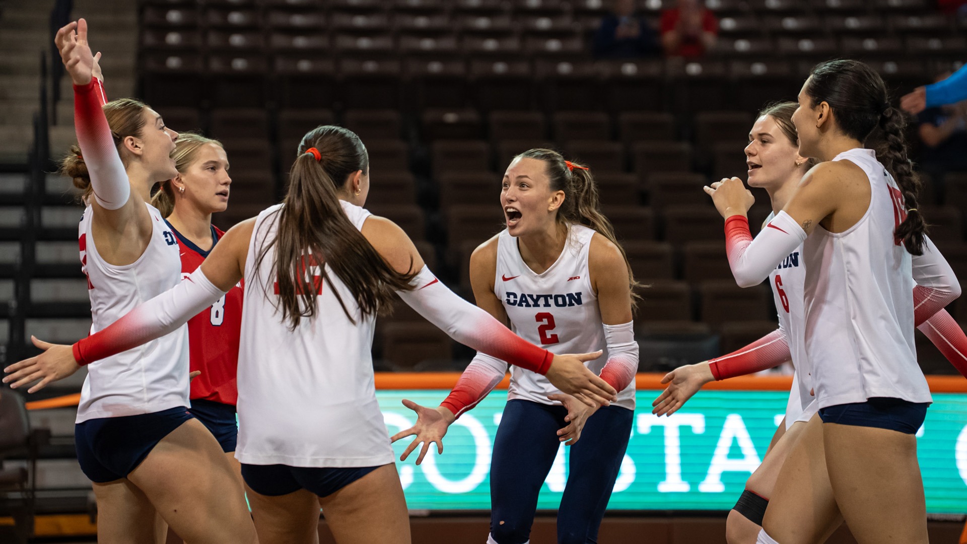 Dayton players celebrate on the court after a point. They are wearing white jerseys with navy shorts and pants.