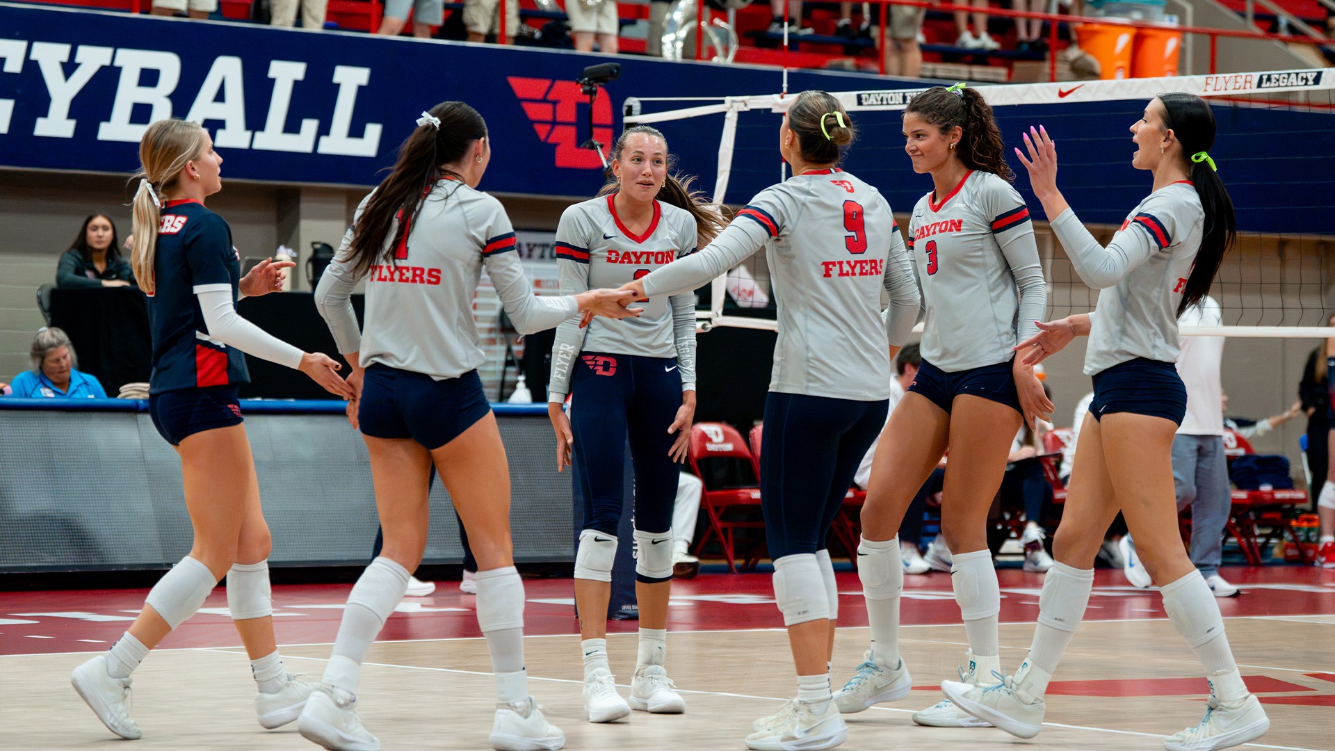 Volleyball players begin to huddle on the court after a point. They are wearing grey jerseys with navy bottoms.
