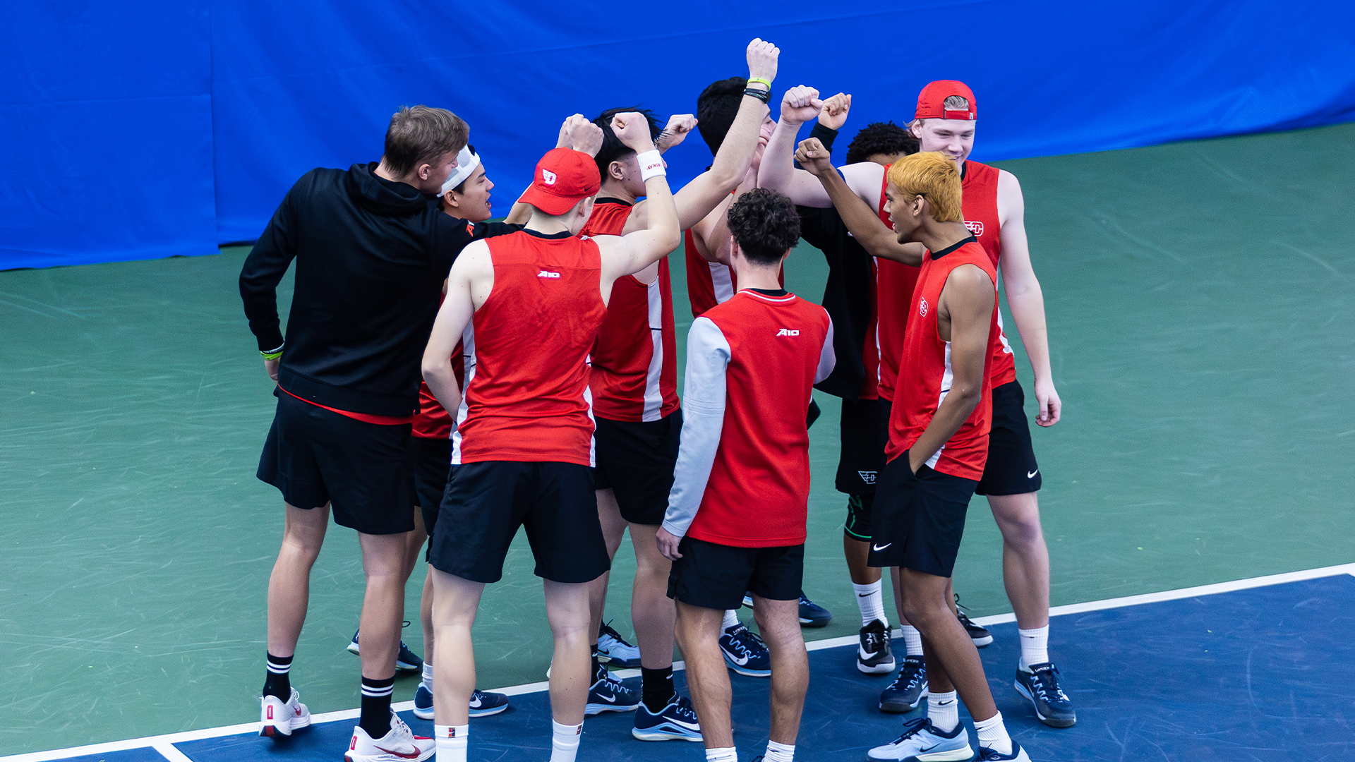 The men's tennis team huddles before a match. Their fists are raised in the middle of the huddle.