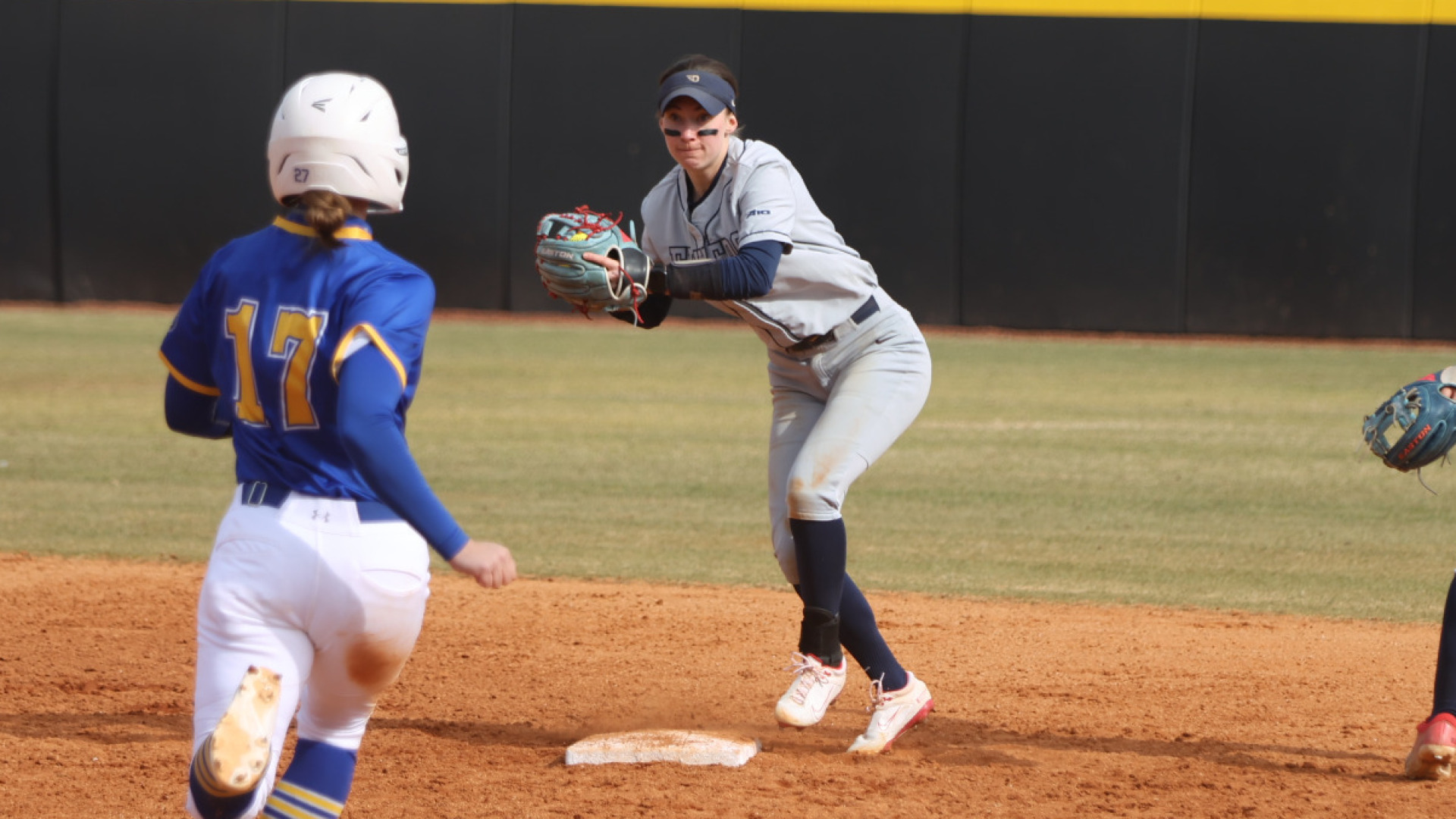 Softball short stop Kirnan Bailey, wearing a gray uniform with blue accents, blue knee-high socks and a blur visor hat, prepares to throw the base to the first baseman from second base during a defensive play in the infield against Morehead State