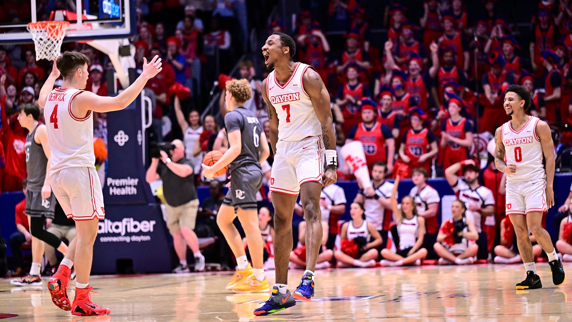 Dayton’s Keonte Jones, wearing No. 7, celebrates with a roar near the basket at UD Arena after a big play, turning toward teammate No. 4 for a high-five while another Flyer, No. 0 Javon Bennett, smiles nearby, with the red-clad crowd and cheerleaders reacting in the background.