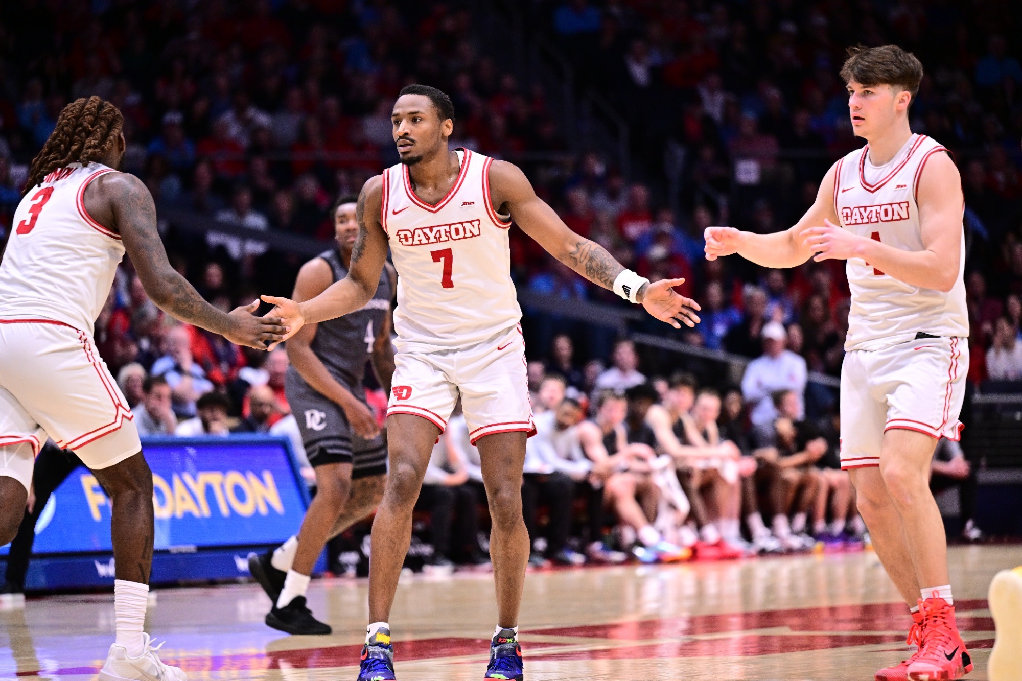 Dayton men’s basketball player Keonte Jones in a white “DAYTON” jersey extends his arms while exchanging a low handshake with a teammate during live game action at UD Arena. The crowd fills the arena seats behind them.