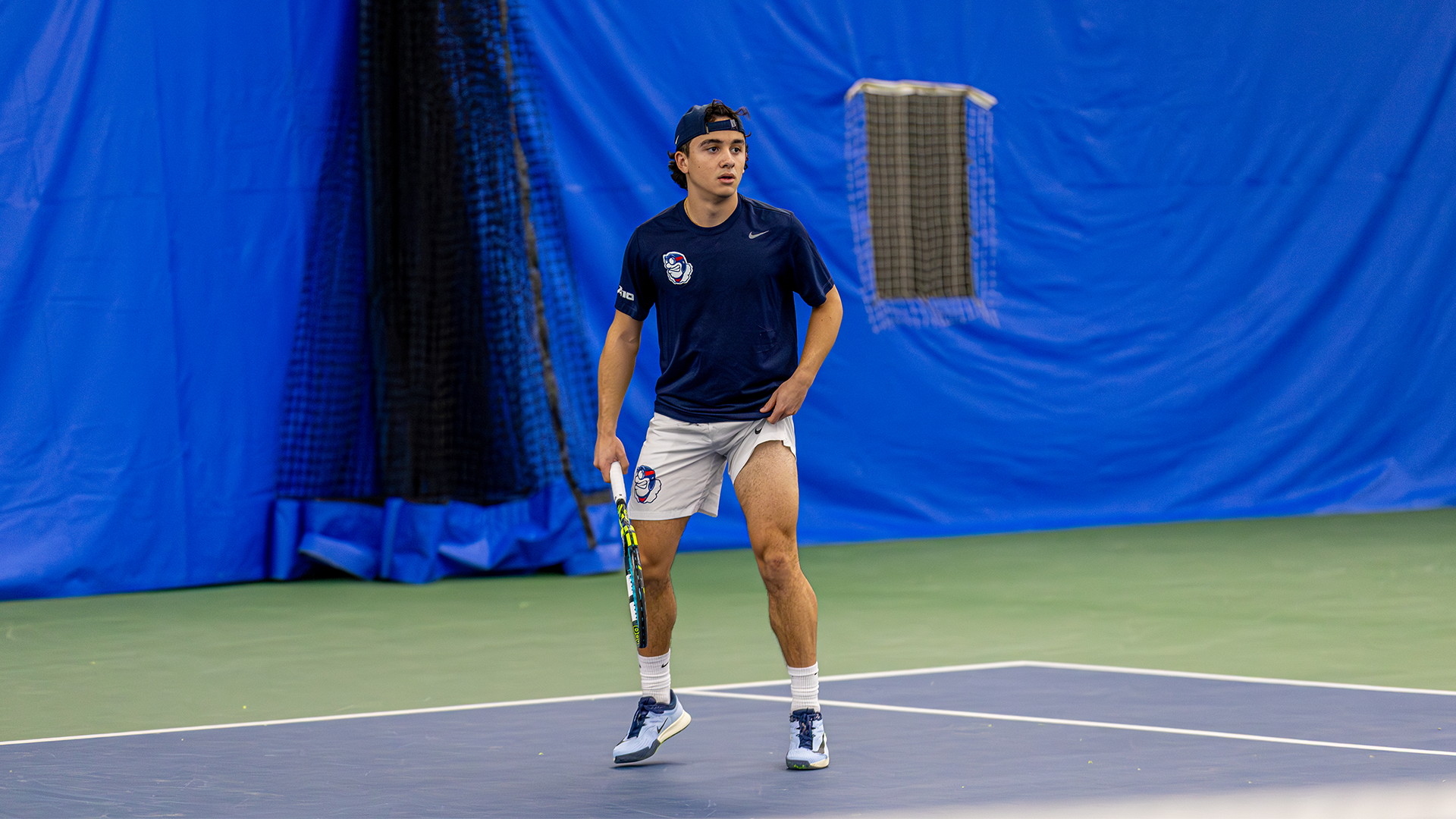 Dylan Jaen bounces on the balls of his feet, ready for the serve. He has a hat on backwards and has his racquet in his right hand.