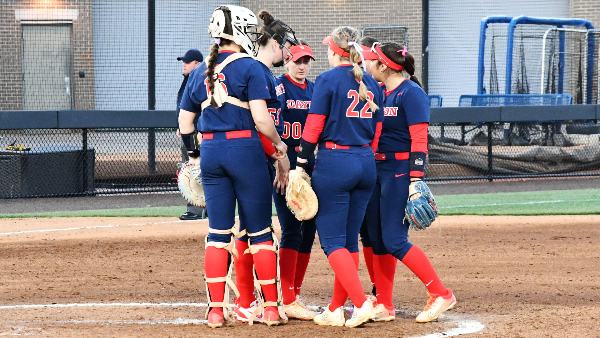 Members of the Dayton softball infield, wearings blue uniforms with red numbers, huddle together near the circle during a game at Kentucky