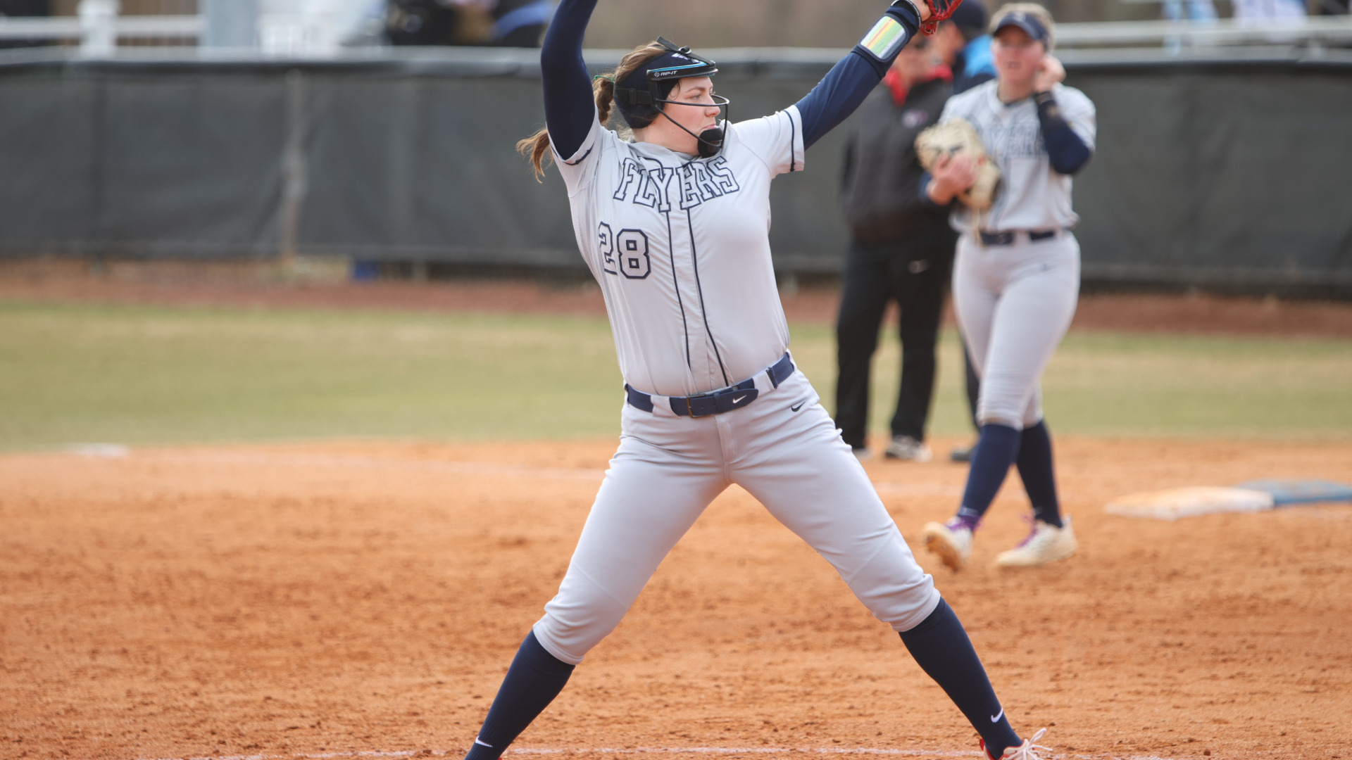 Pitcher Kate Kastelic with her hand above her head in the middle of a pitch delivery wearing gray top and bottom uniform during a game PC Softball Stadium