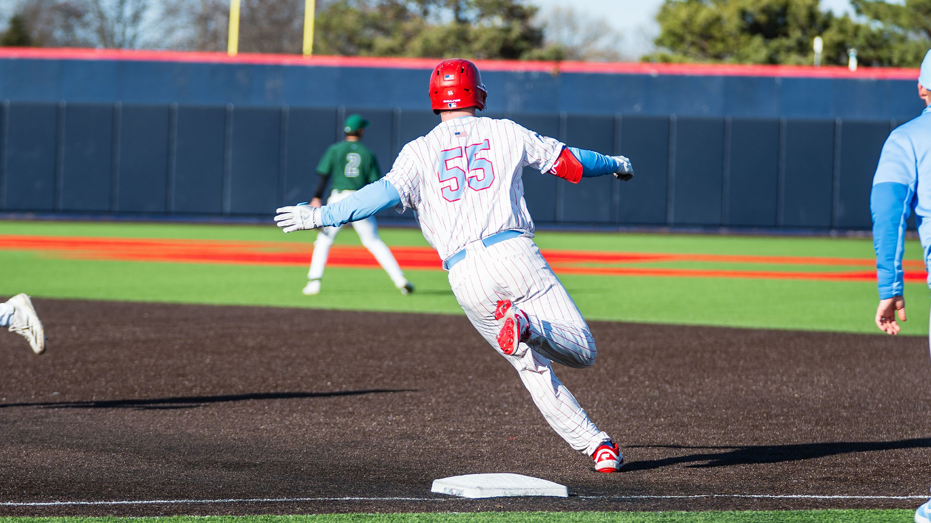 Ryan MacDougall rounds first base. He is wearing a white uniform with red pinstripes and a red helmet. His back is to the camera.
