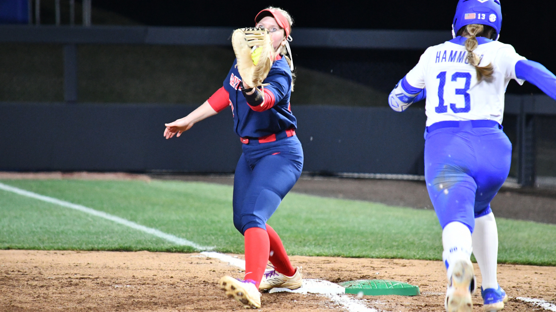 Softball's #22 Molly Grace, wearing a blue uniform top and bottom, catches the ball at first base ahead of a Kentucky base runner wearing a white uniform top and blue uniform pants.