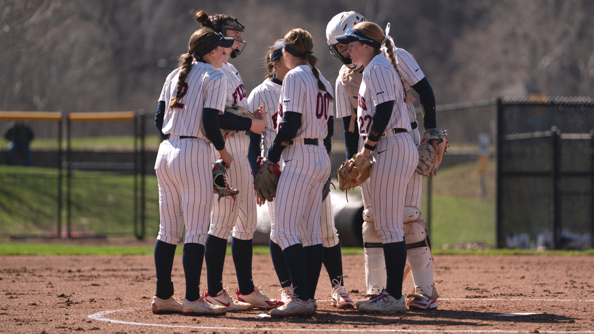 Softball infielders, wearing white pinstripe uniforms, gather inside the circle before the start of a game against Loyola Chicago at UD Softball Stadium