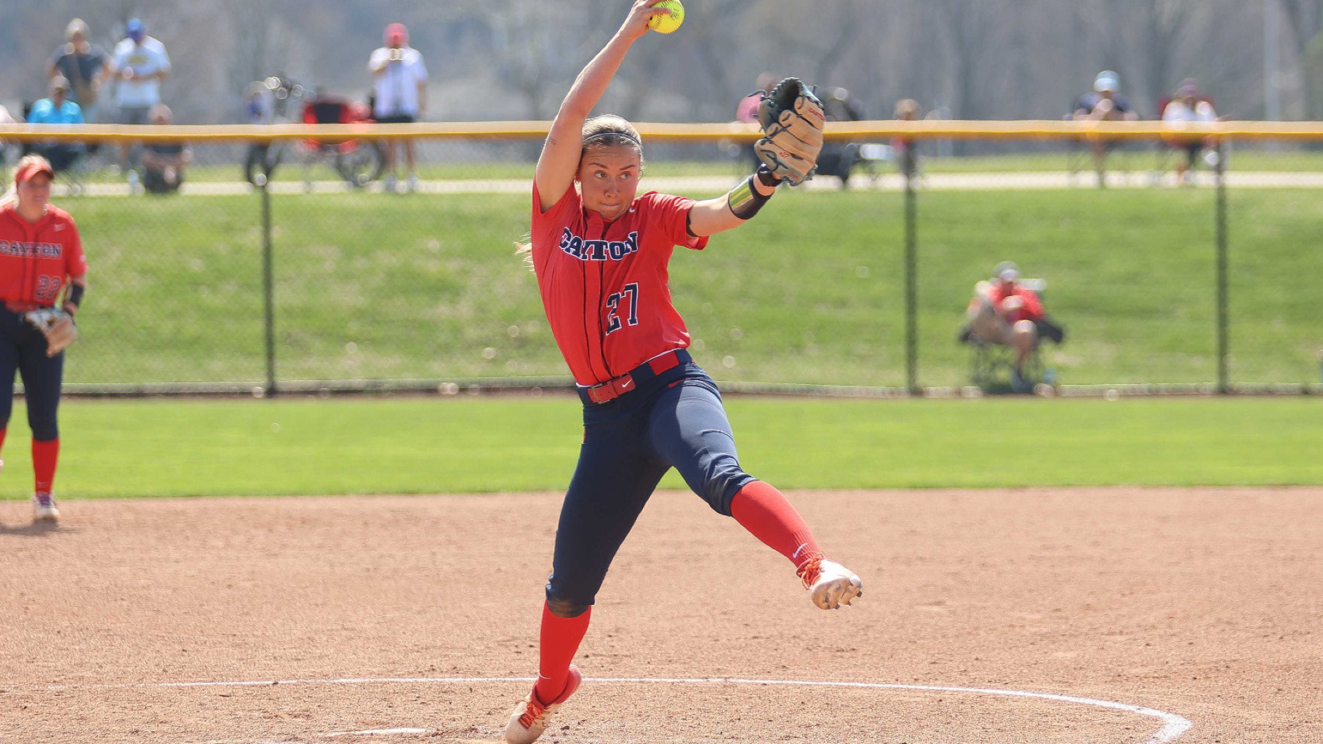 Softball pitcher Camryn Craig, wearing a red Dayton uniform top with the number 27 and blue pants, with the ball above her head in the top of her throwing motion inside the circle during a game at UD Softball Stadium