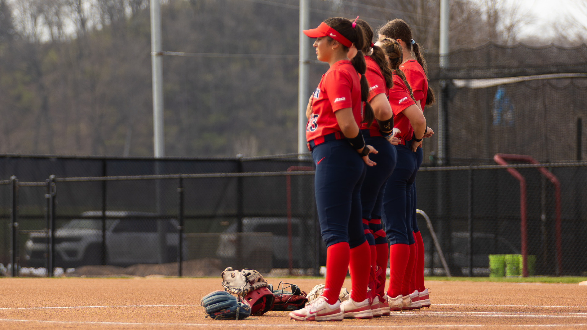 Four members of the softball team, wearing red uniform tops and blue pants, standing in the circle facing the flag during the national anthem