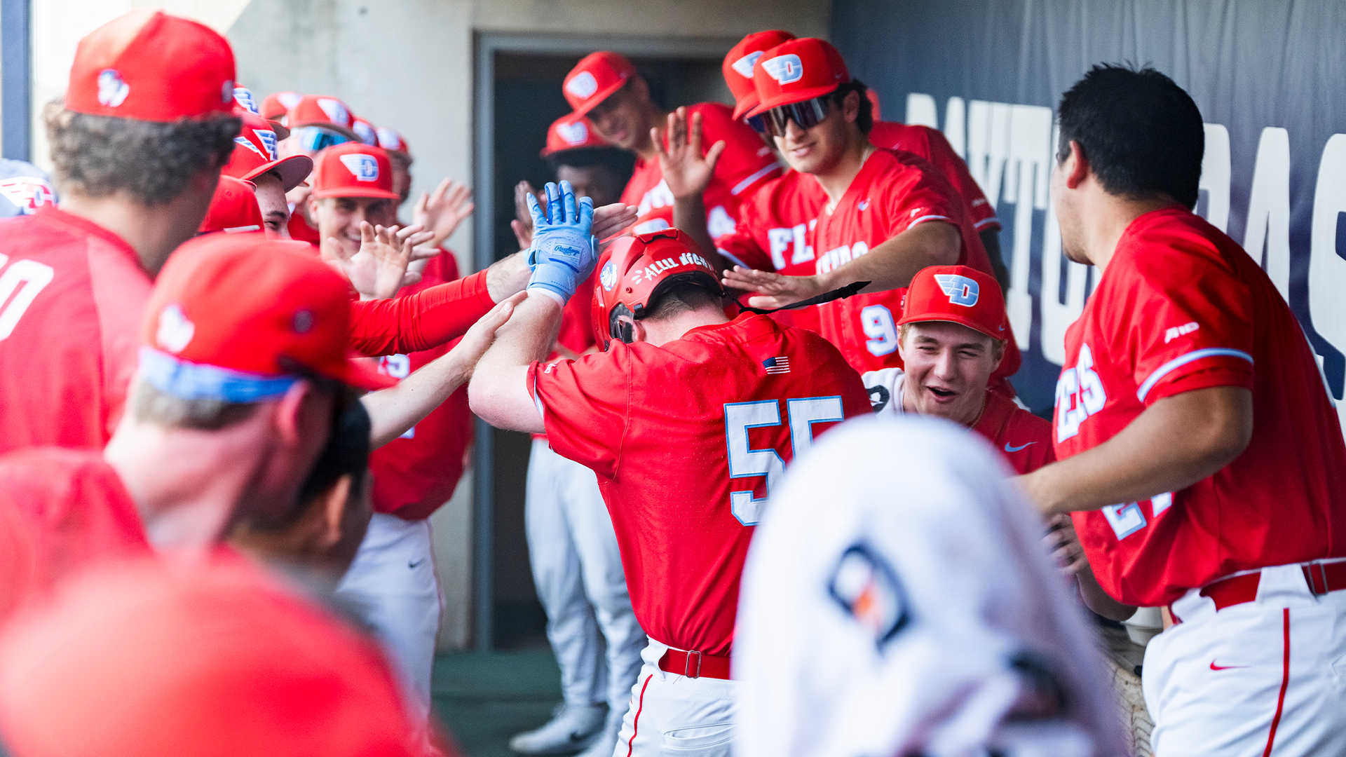 Ryan MacDougall celebrates with teammates in the dugout. He is wearing a red helmet, while other players smack his helmet.