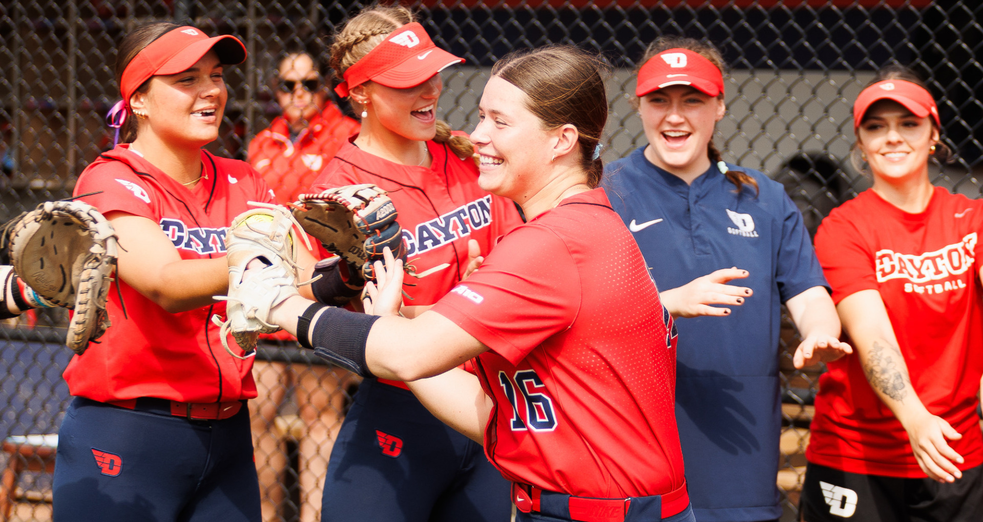 Members of the softball team, wearing red uniform tops and blus pants, celebrate during starting lineups as number 16 slaps hands and faces toward the field