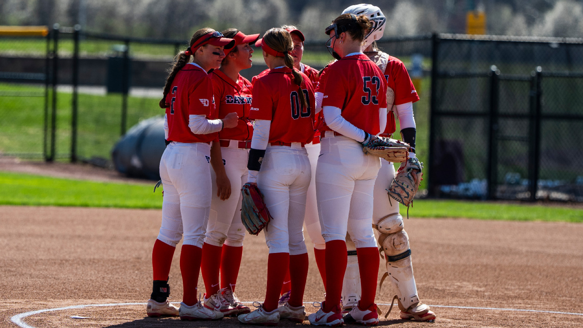 Members of the softball infield, wearing red uniform tops and blue pants, gather in the circle with the pitcher and the catcher, during a game at UD Softball Stadium