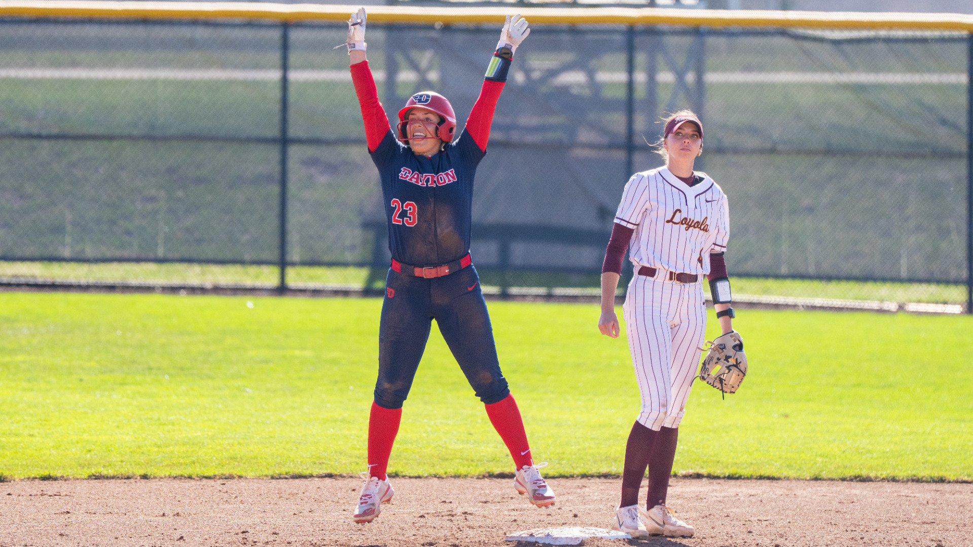 Softball's number 23 Juliana Gonzalez, wearing a blue uniform top and pants with red sleeves and socks with a red batting helmet, jumps with two hands in the air at second base to celebrate hitting a double