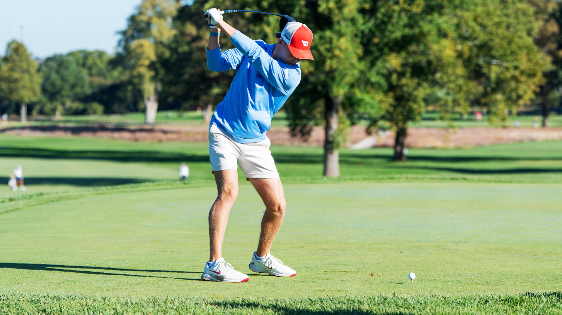 Men's golfer TJ Kreusch, wearing a light blue long-sleeved top, khaki shorts and a red and white trucker hat, at the top of his back swing in a tee box at Moraine Country Club