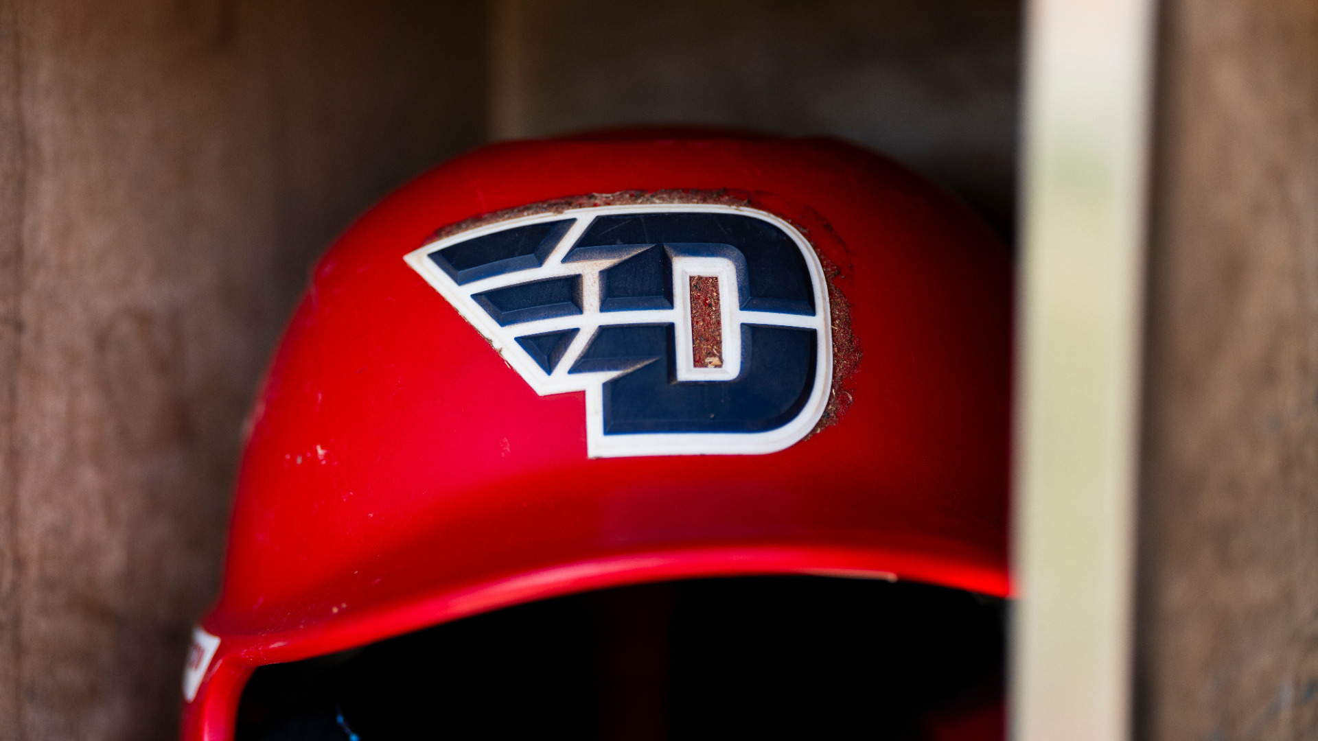 A red softball batting helmet with a blue D logo sitting inside a cubby in the dugout 