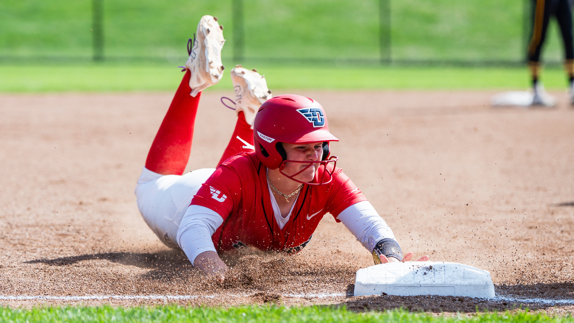 Softball's Molly Grace, wearing a red jersey top, white pants and a red batting helmet, slides into third base during a game at UD Softball Stadium