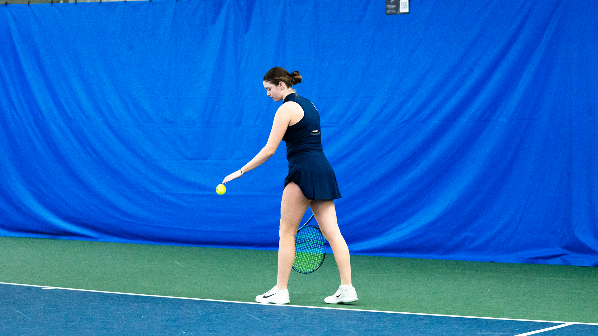 Baby Jordan, wearing a navy uniform, bounces a ball on the ground prior to serving.