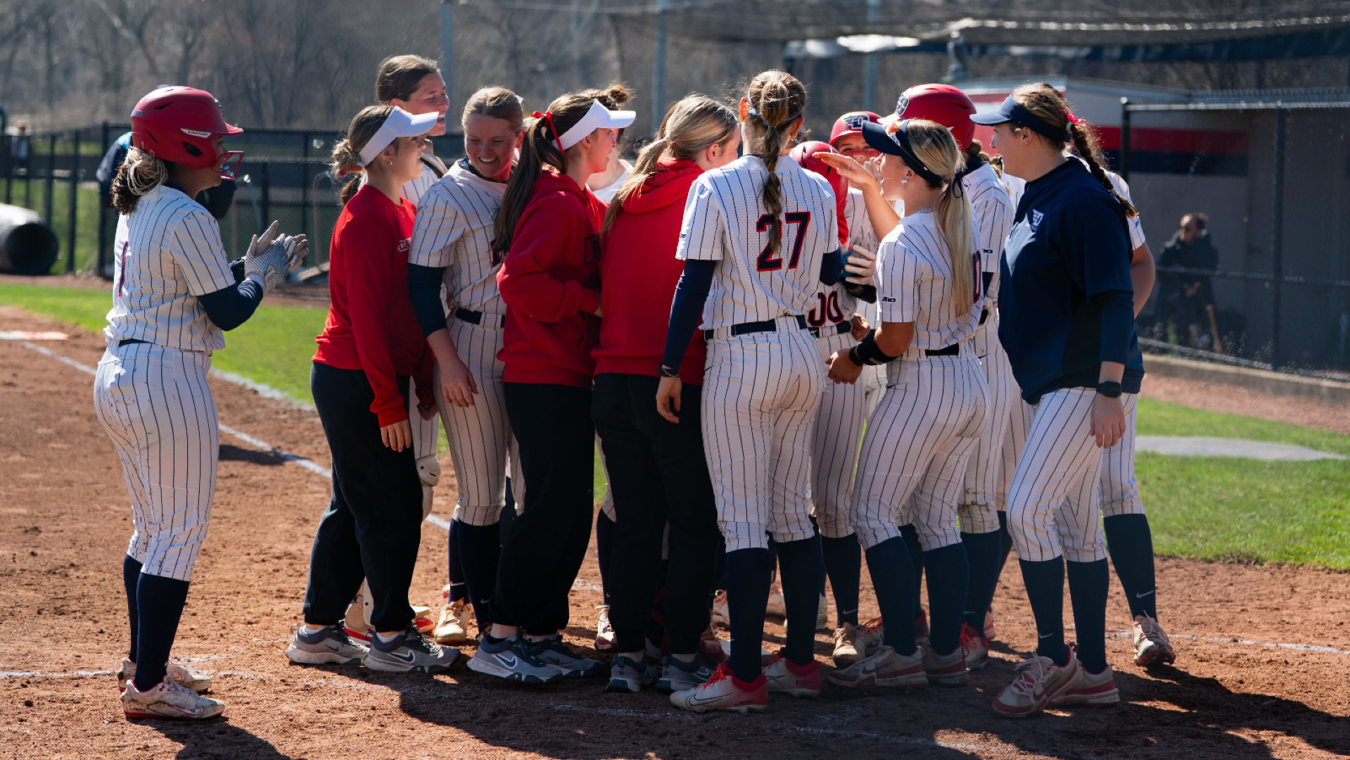 Members of the Dayton softball team, wearing white pin stripe uniforms, gather around home plate to celebrate a home run at UD Softball Stadium