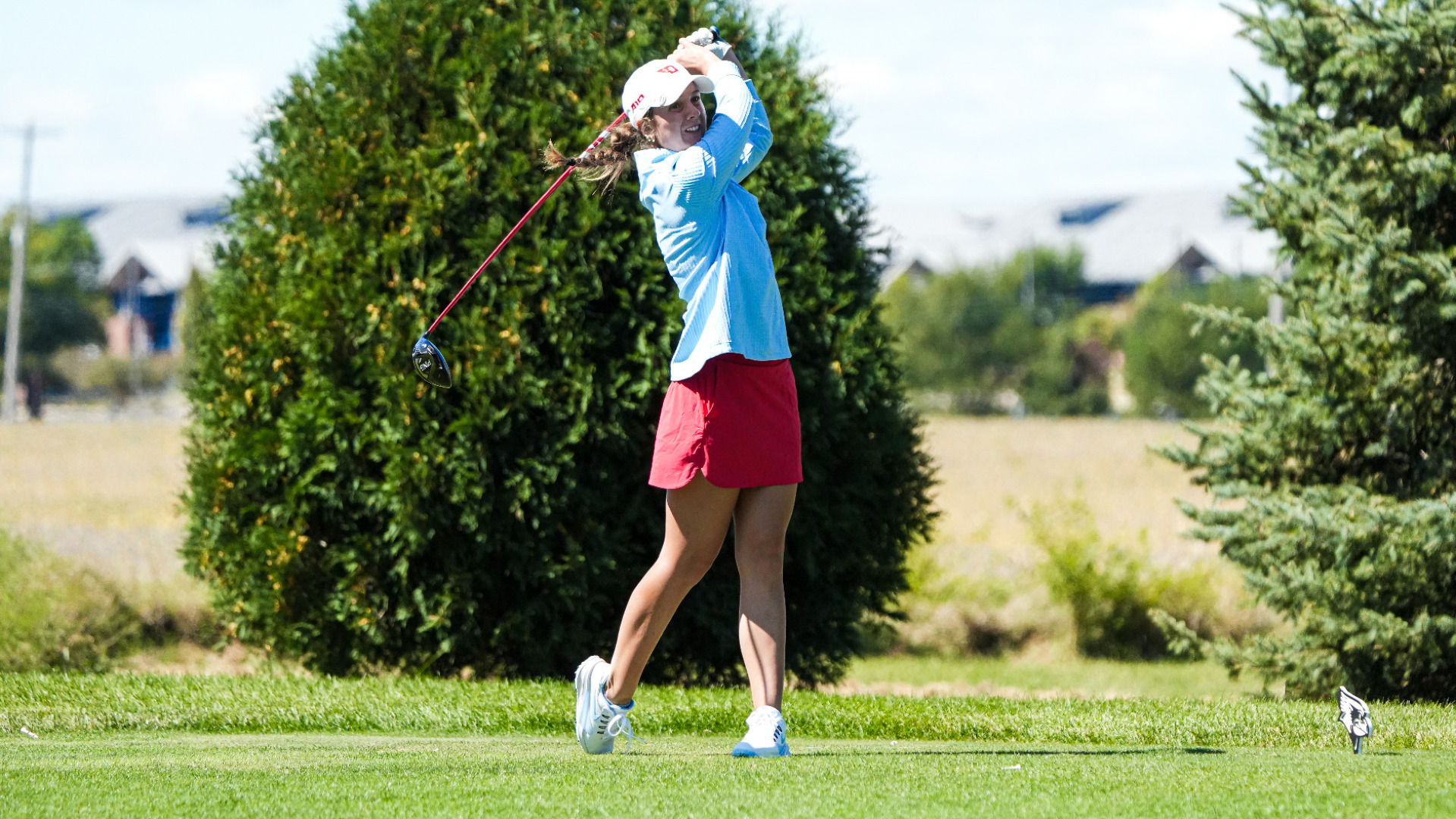 Women's golfer Emma Sparling, wearing a light blue long sleeve top, red skirt and white hat, in her back swing on a tee box at ISU's Redbird Invitational