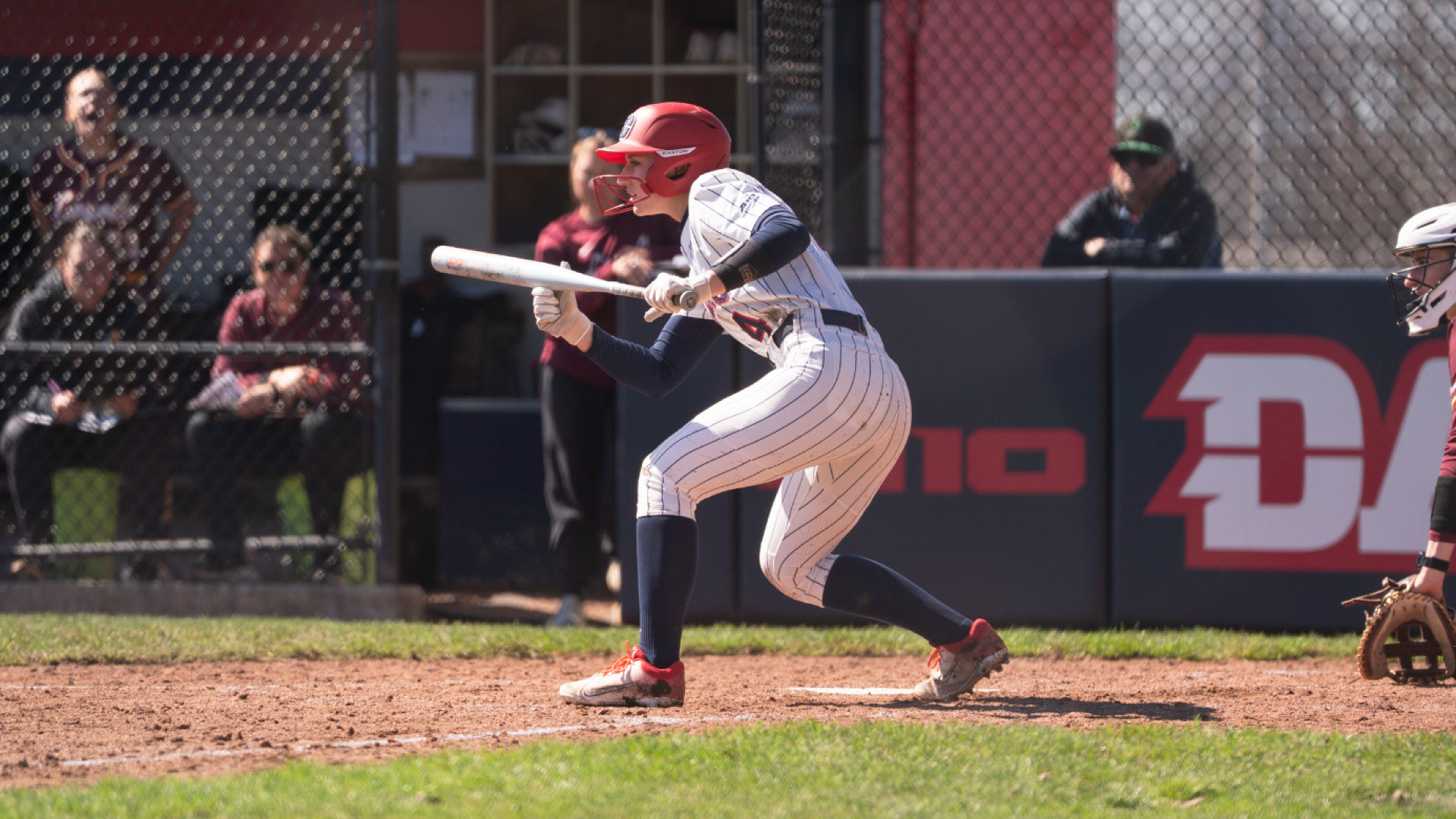 Softball's number 4 Mickie Shively, wearing a white pinstripe uniform with blue socks and a red batting helmet, squares to bunt in the batter's box during a game at UD Softball Stadium