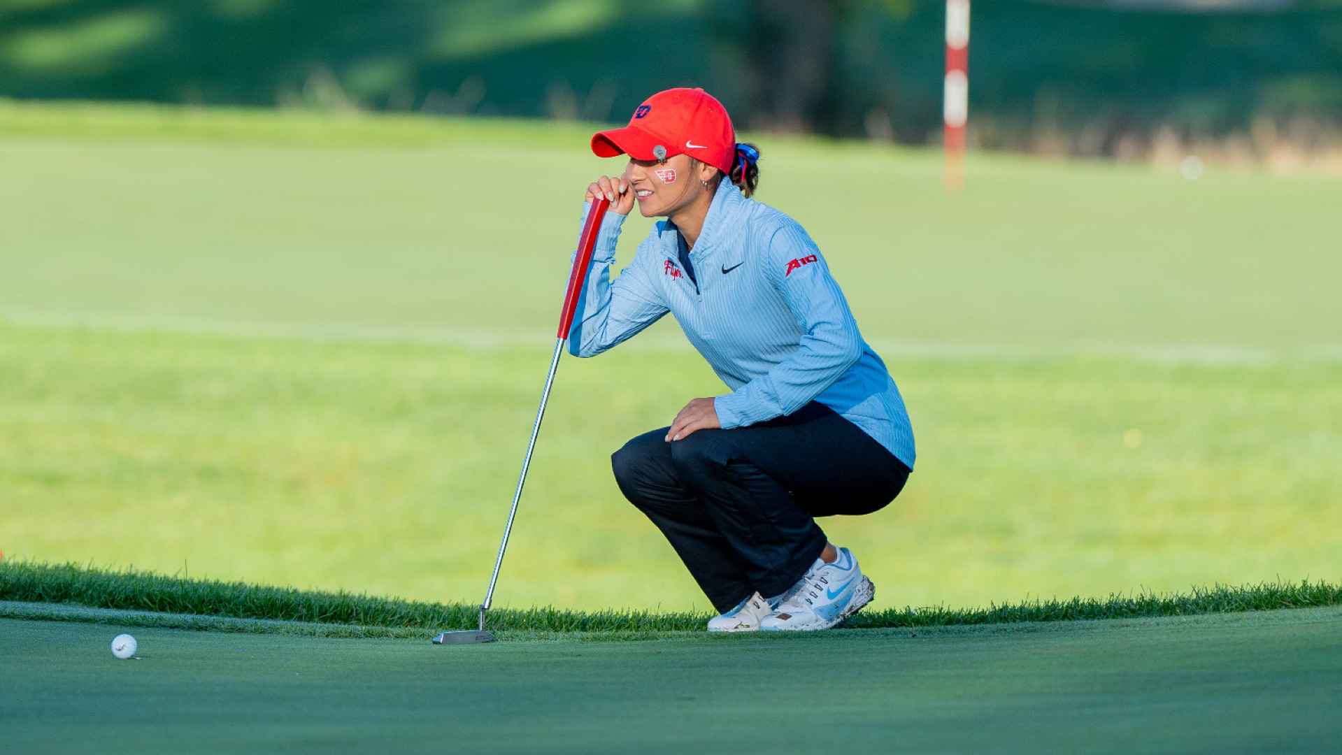 Women's golfer Christina Pfefferkorn, wearing a light blue pull over, navy pants and a red hat, crouches down on a green with her putter in her hand to check the lie of the green