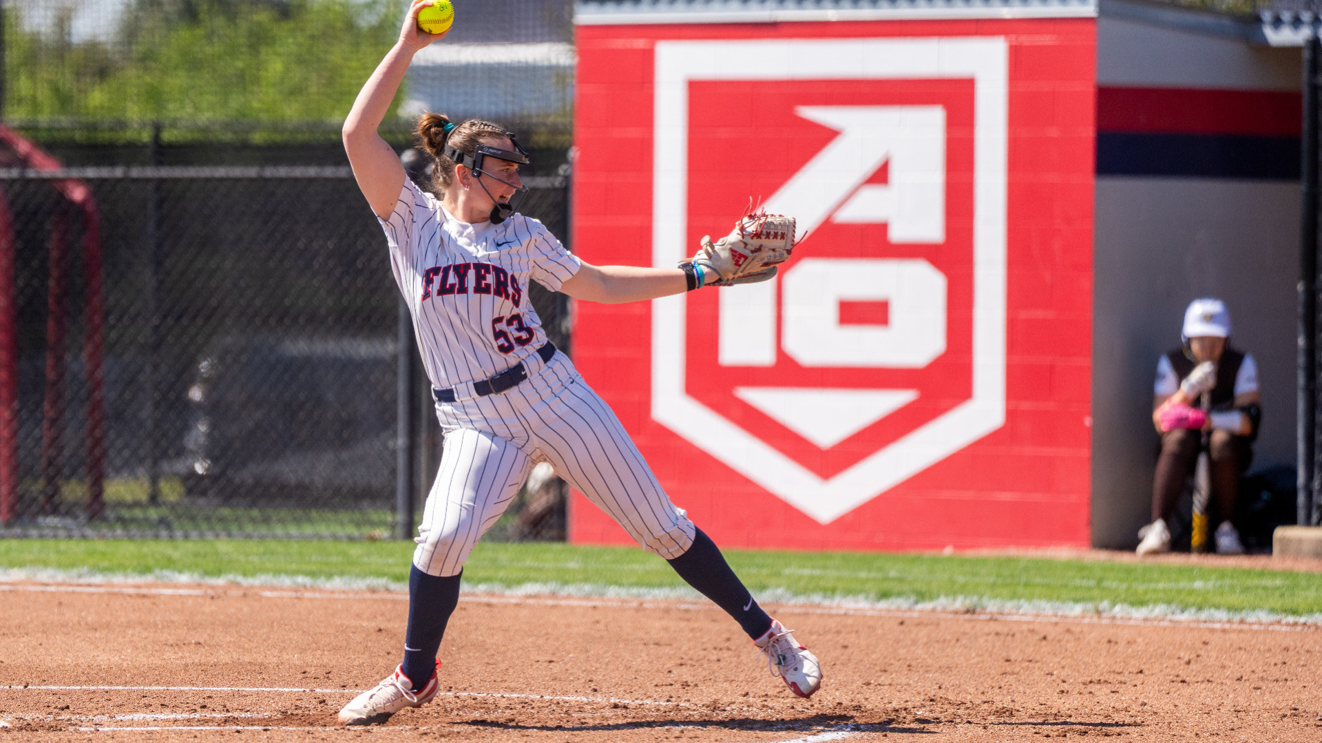 Softball player Izzy Kemp, wearing a white pinstripe uniform with the word Flyers across the chest and the number 53, is in the pitcher's circle with a ball above her head in the top of her throwing motion during a game at UD Softball Stadium
