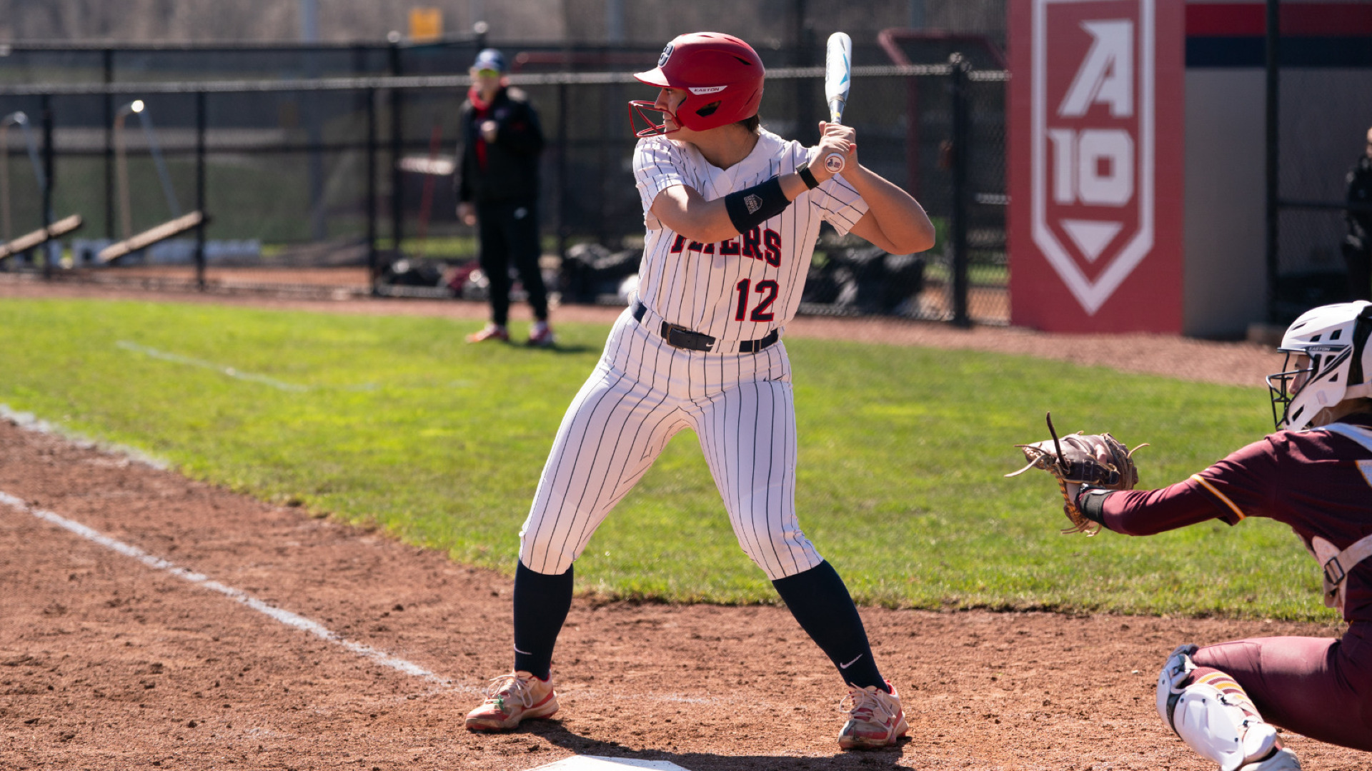 Soseh Broglin, wearing a white pin stripe uniform with the number 12 and Flyers across the chest along with a red batting helmet, stands in the batter's box with her bat on her shoulder awaiting a pitch during a game at UD Softball Stadium