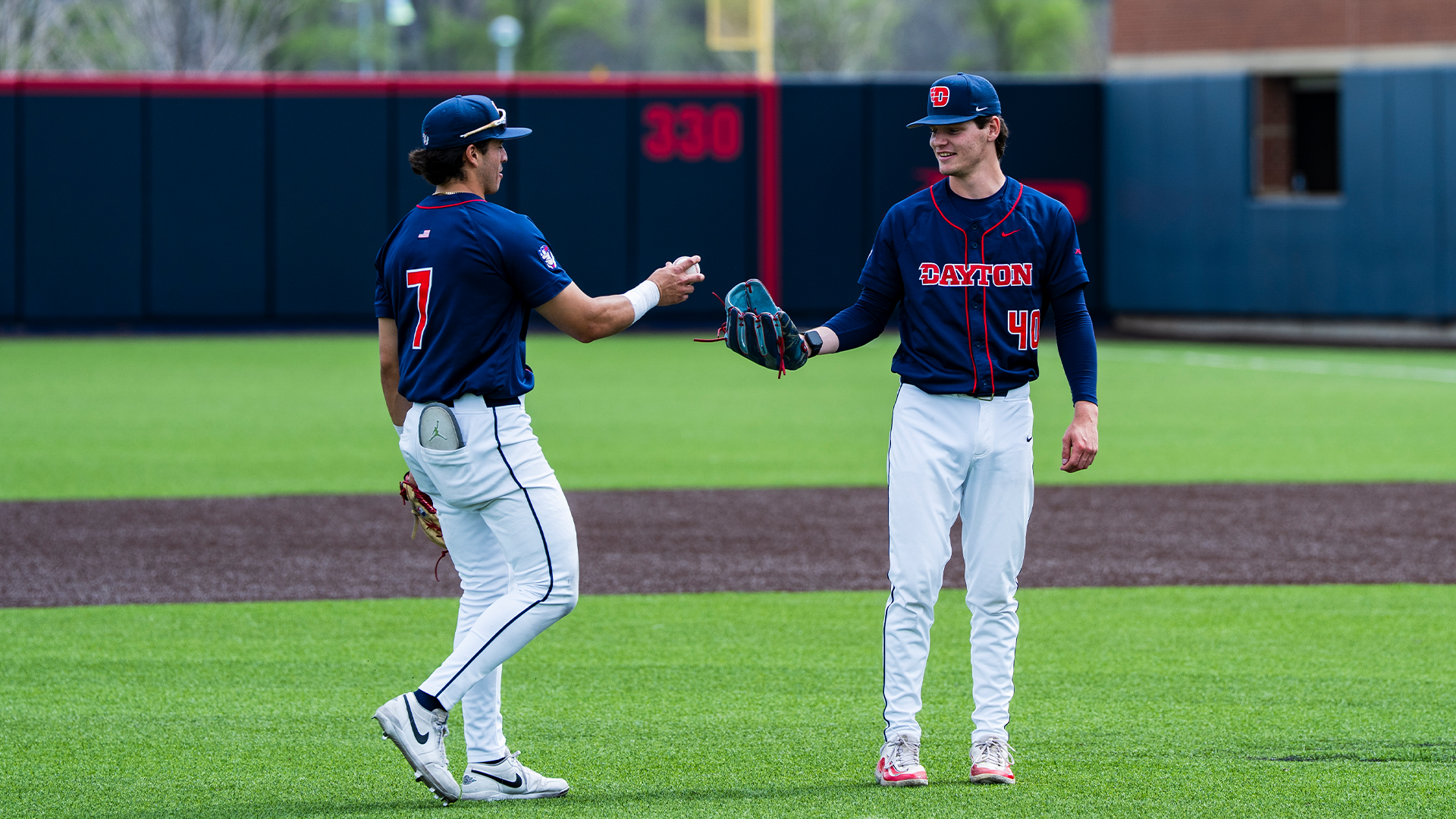 Jason Bello puts a baseball into Carson Samuels' glove. Both players are in navy jerseys, white pants and a navy hat.