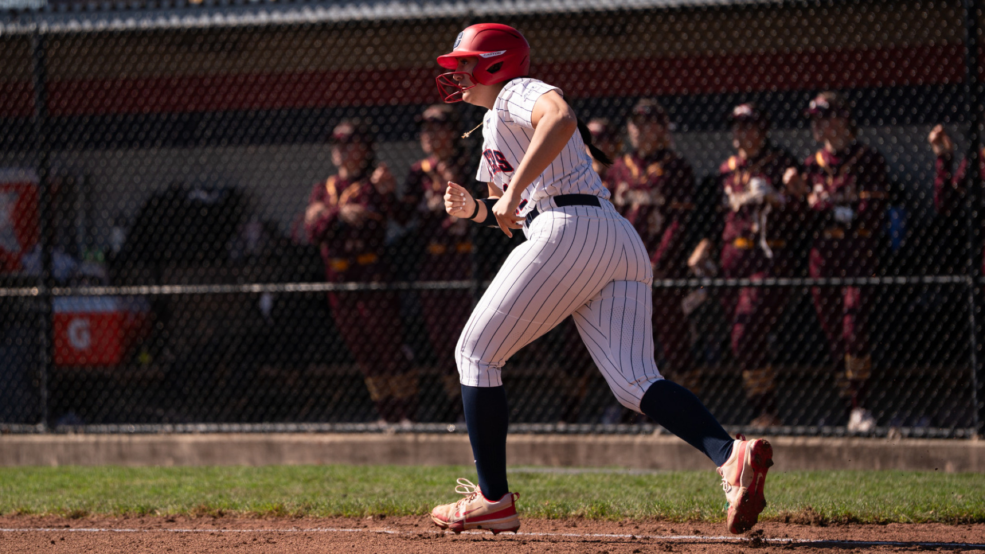 Softball's number 12, wearing a white pinstripe uniform and a red batting helmet, takes off running down the first base line, in front of the visiting team dugout, after a hit during a game at UD Softball Stadium