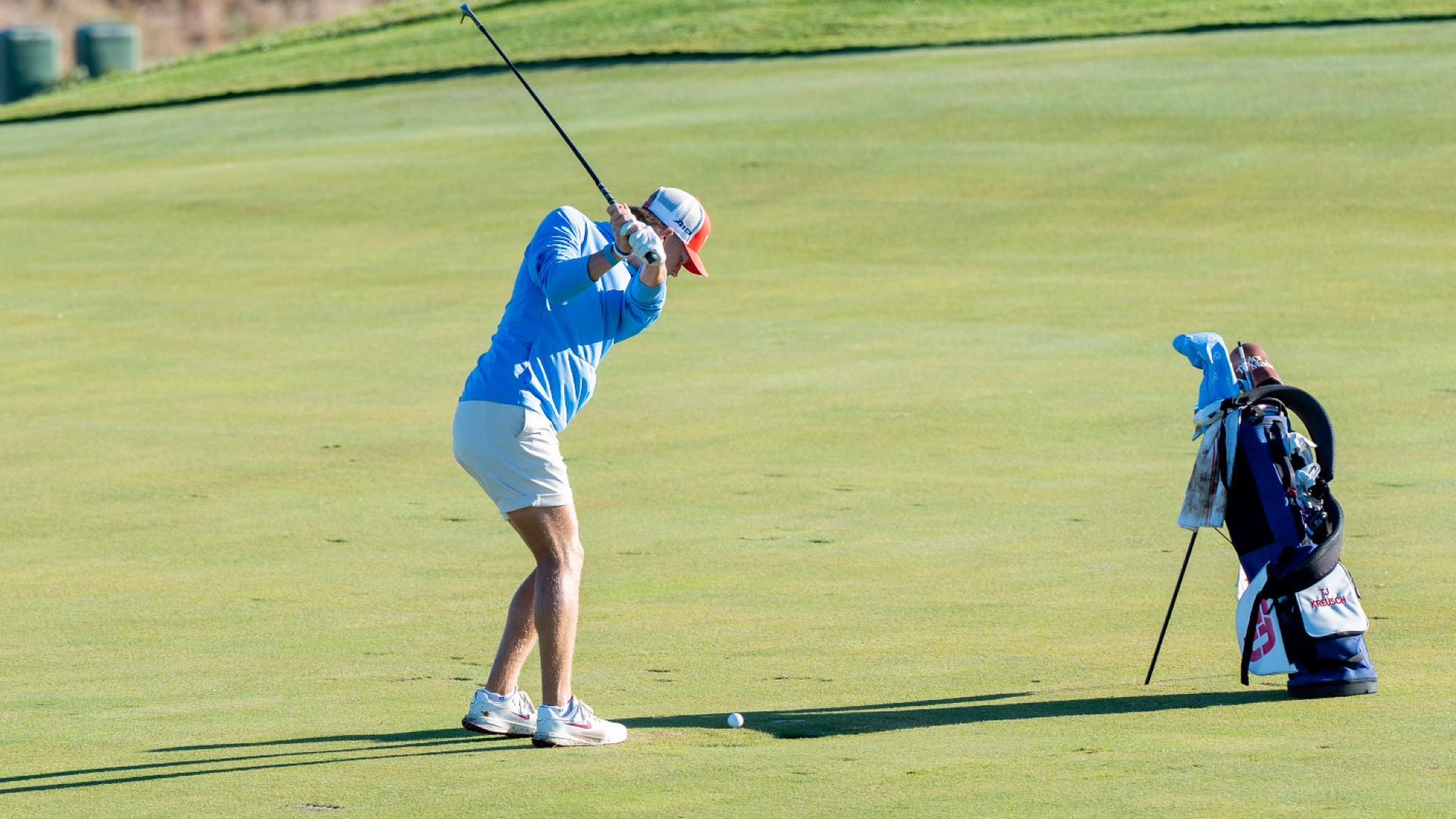 Men's golfer T.J. Kreusch, wearing a long-sleeve light blue top, khakie shorts and a white and red trucker-style hat, is in the top of his back swing in a fairway at Moraine Country Club. His golf bag is in front of him