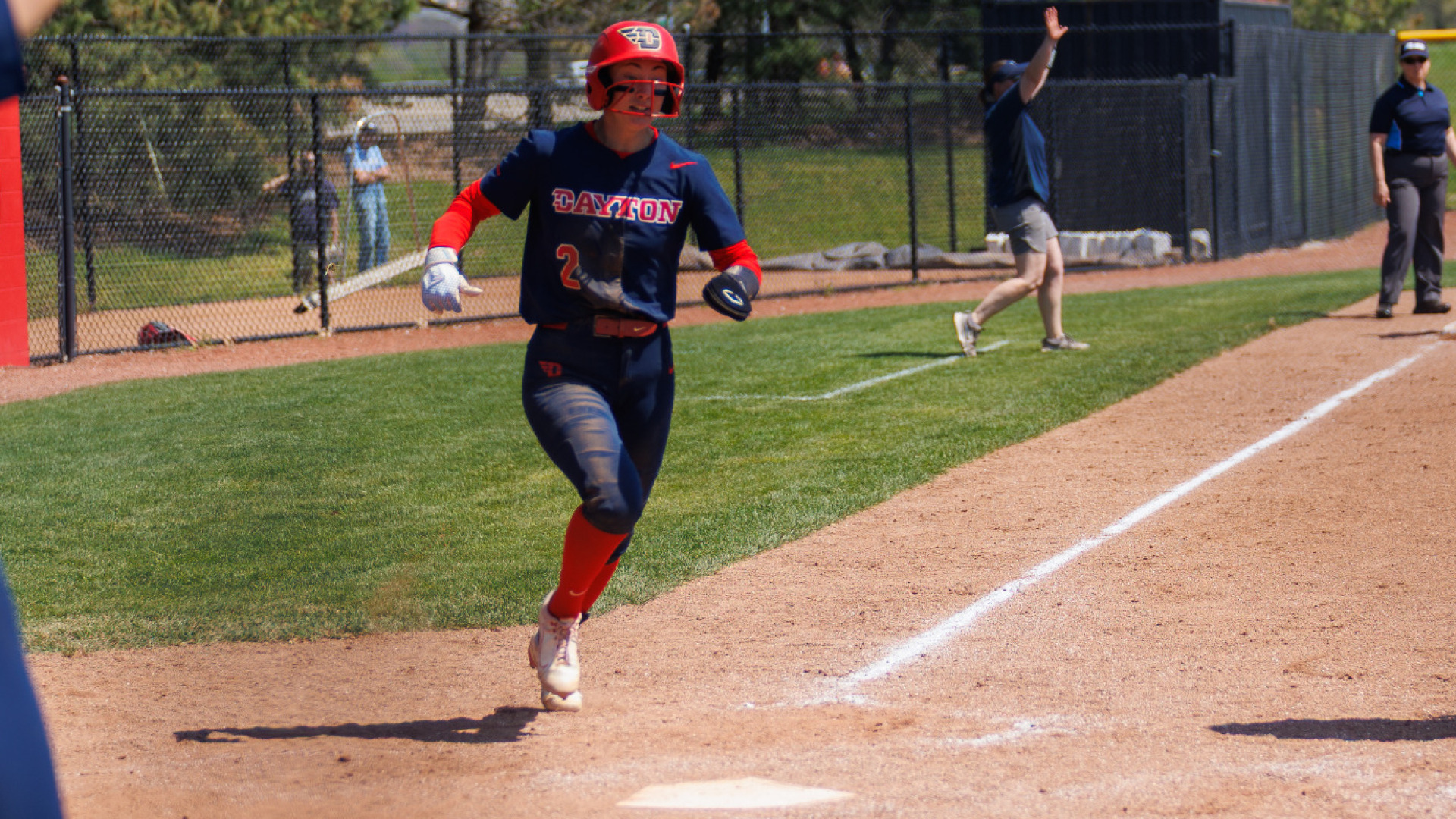 Softball's number two, wearing a blue uniform top and bottom, black sliding glove o nher left hand and a red batting helmet, runs toward home plate just ahead of scoring a run during a game at UD Softball Stadium