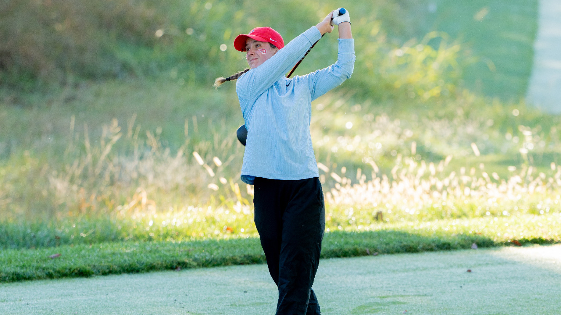 Women's golfer Emma Sparling, wearing a light blue-long-sleeve top, black pants and a red hat, in the top of her backswing in a tee box at NCR Country Club