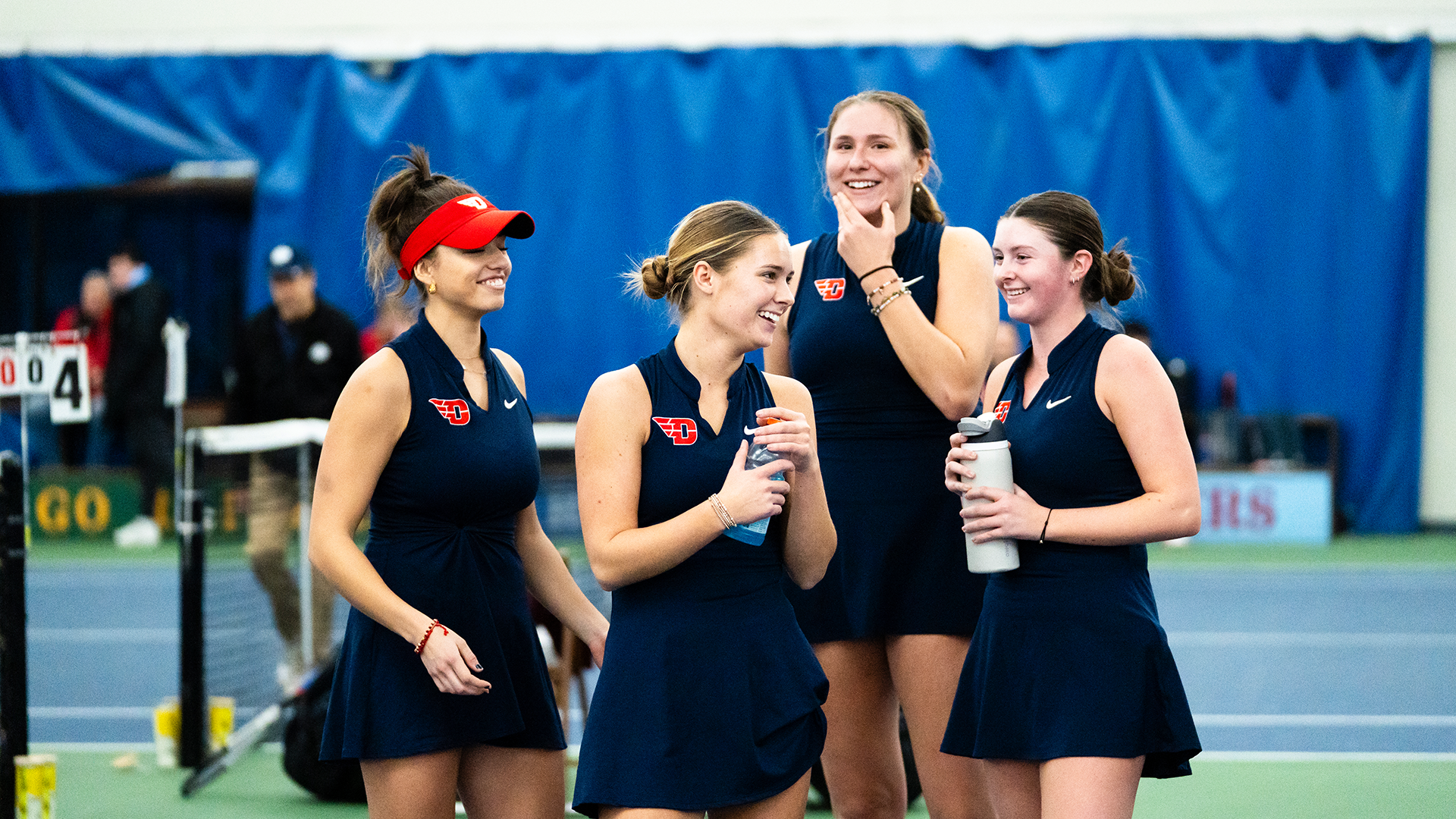 Monserrat Sierra, Mallory Hitchcock, Natalie Osiecki and Baby Jordan stand together smiling while cheering on their teammates.