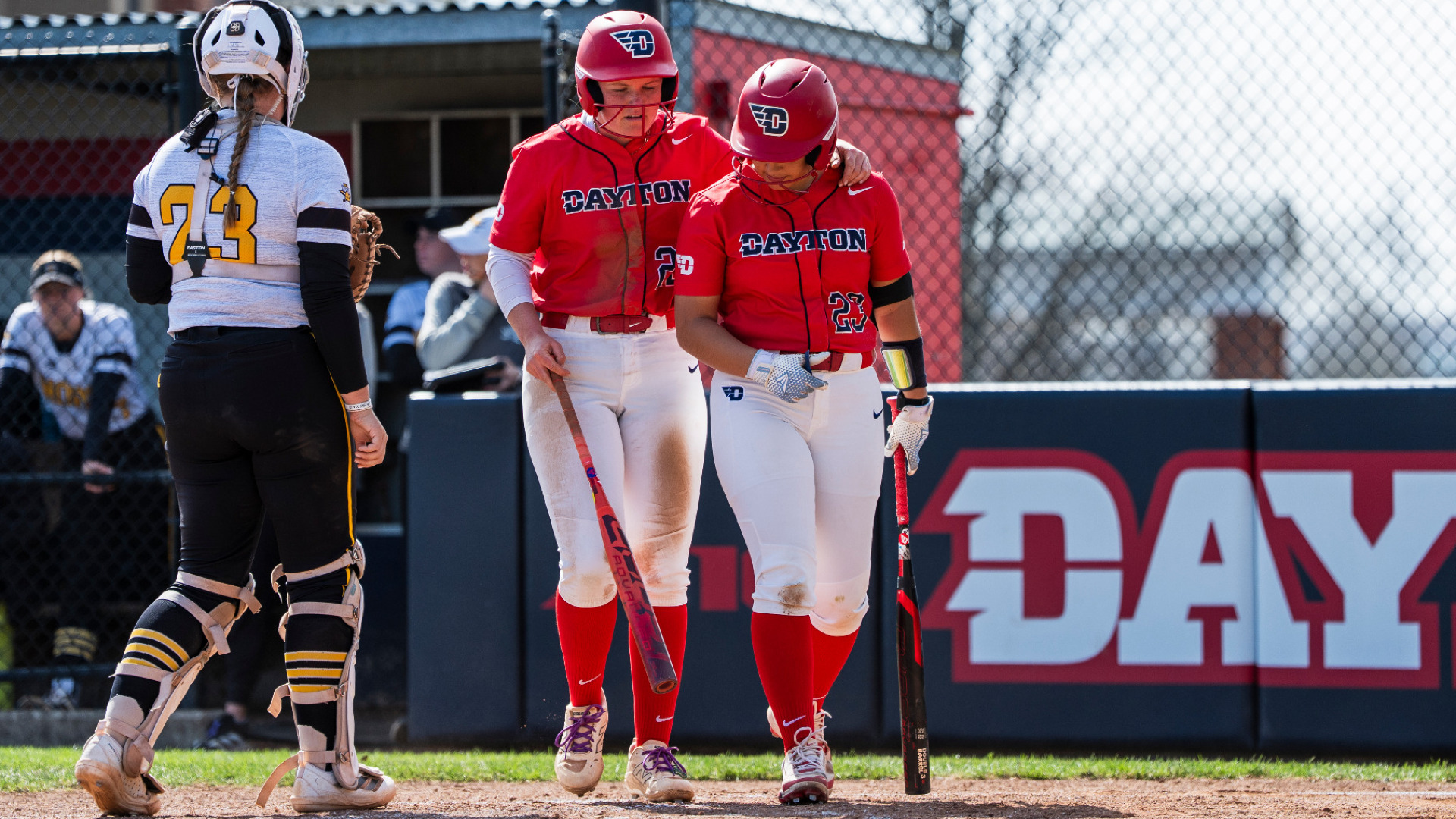 Softball's number 22 Molly Grace (left) and number 23 Juliana Gonzalez (right), wearing red uniform tops, white pants, red socks and red batting helmets, stand side-by-side near homeplate in conversation during a game at UD Softball Stadium